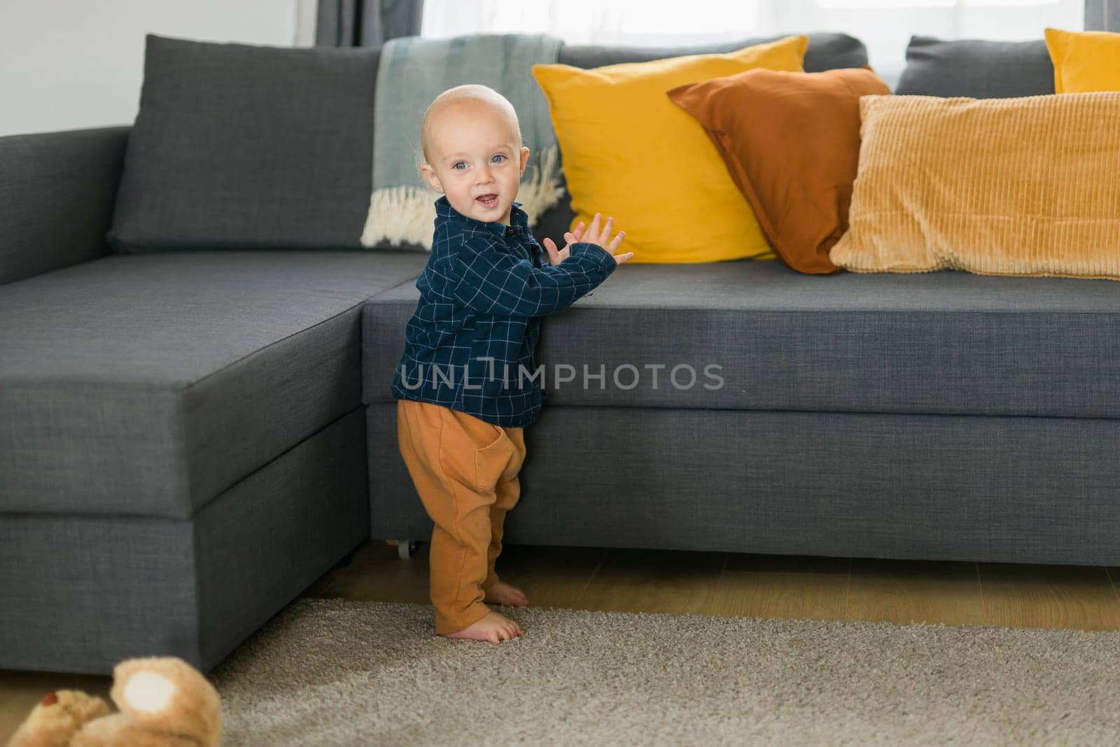Toddler boy laughing having fun standing near sofa in living room at home copy space. Adorable baby making first steps alone. Happy childhood and child care concept by Satura86