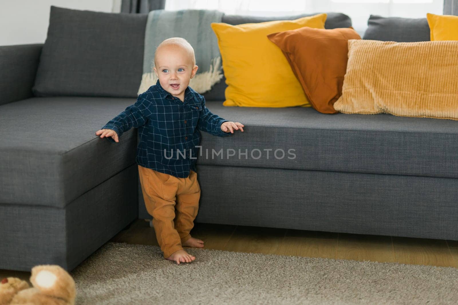 Toddler boy laughing having fun standing near sofa in living room at home. Adorable baby making first steps alone. Happy childhood and child care