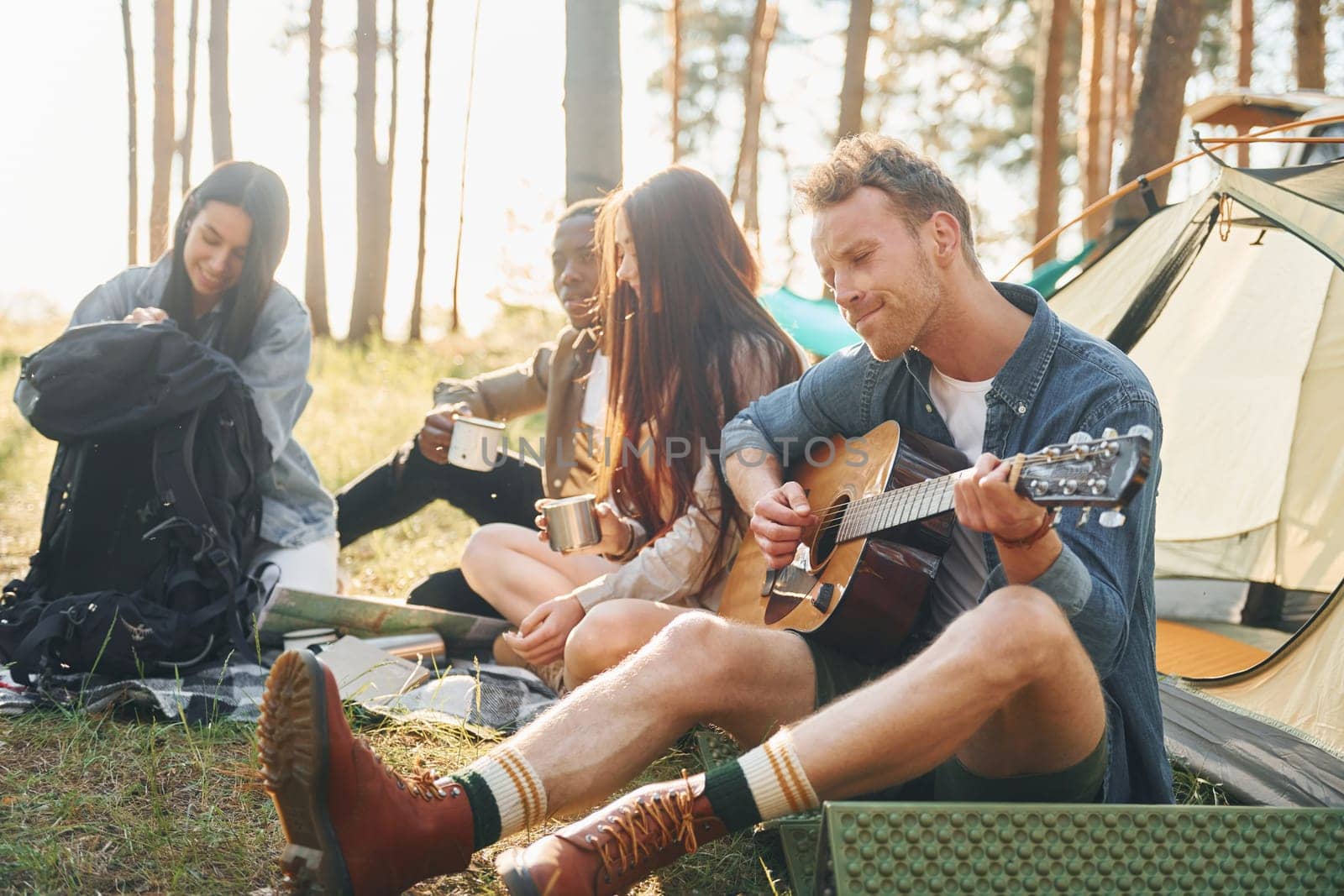 Man plays guitar. Group of young people is traveling together in the forest at daytime.