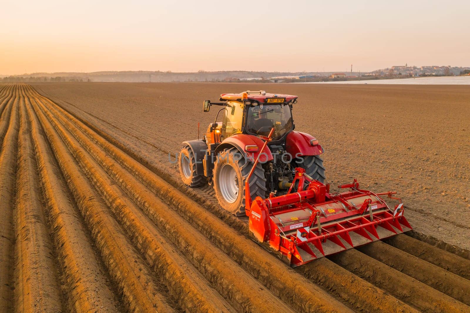 PRAGUE , CZECH REPUBLIC - MARCH 18 2022: Heavy tractor drags plow leaving long furrows behind. Powerful machine tills in field with cultivated soil after sunset at rural site