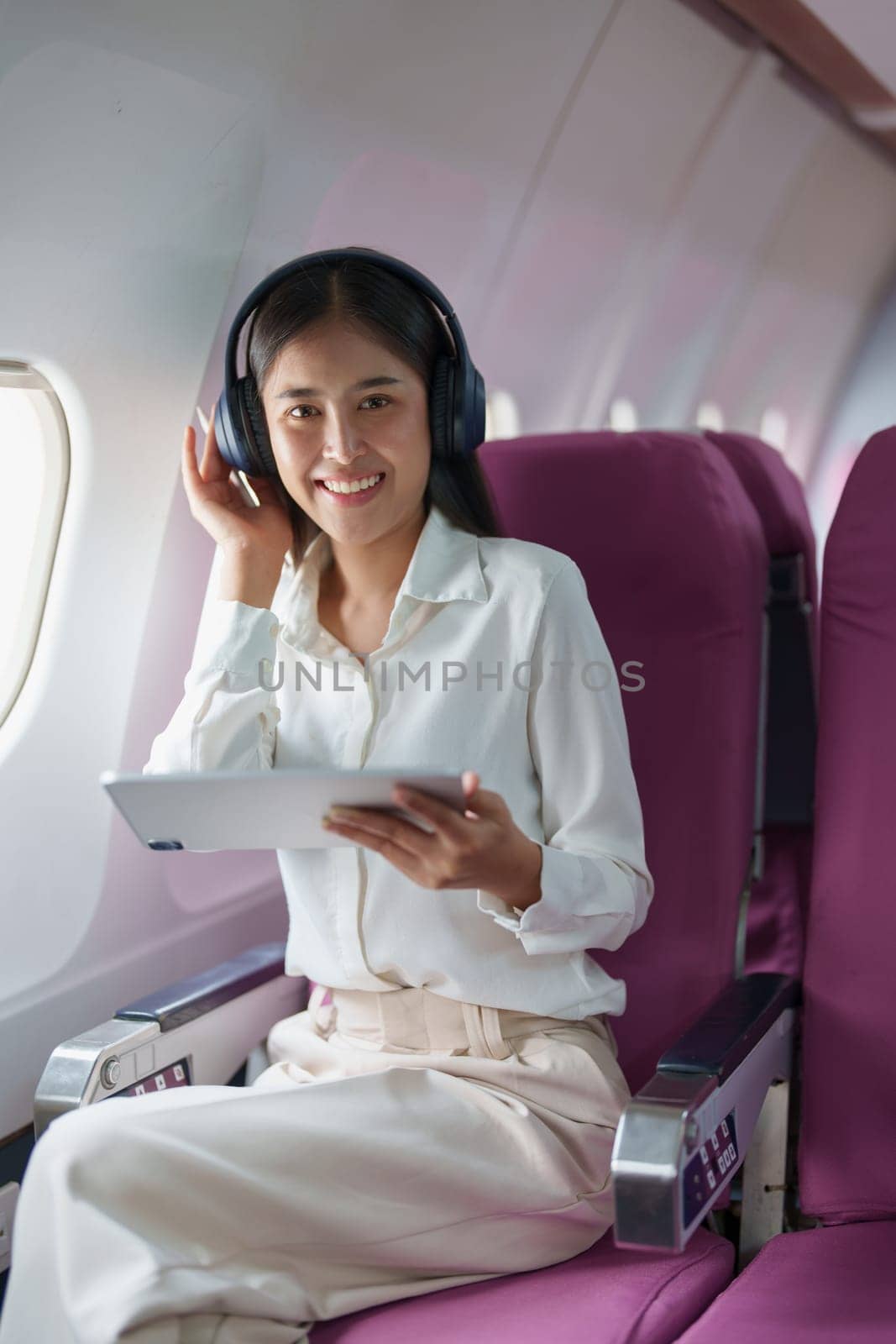 Young Asian attractive business woman passenger sitting on business class luxury plane while wearing wireless headphone for relax and working with tablet during the flight.