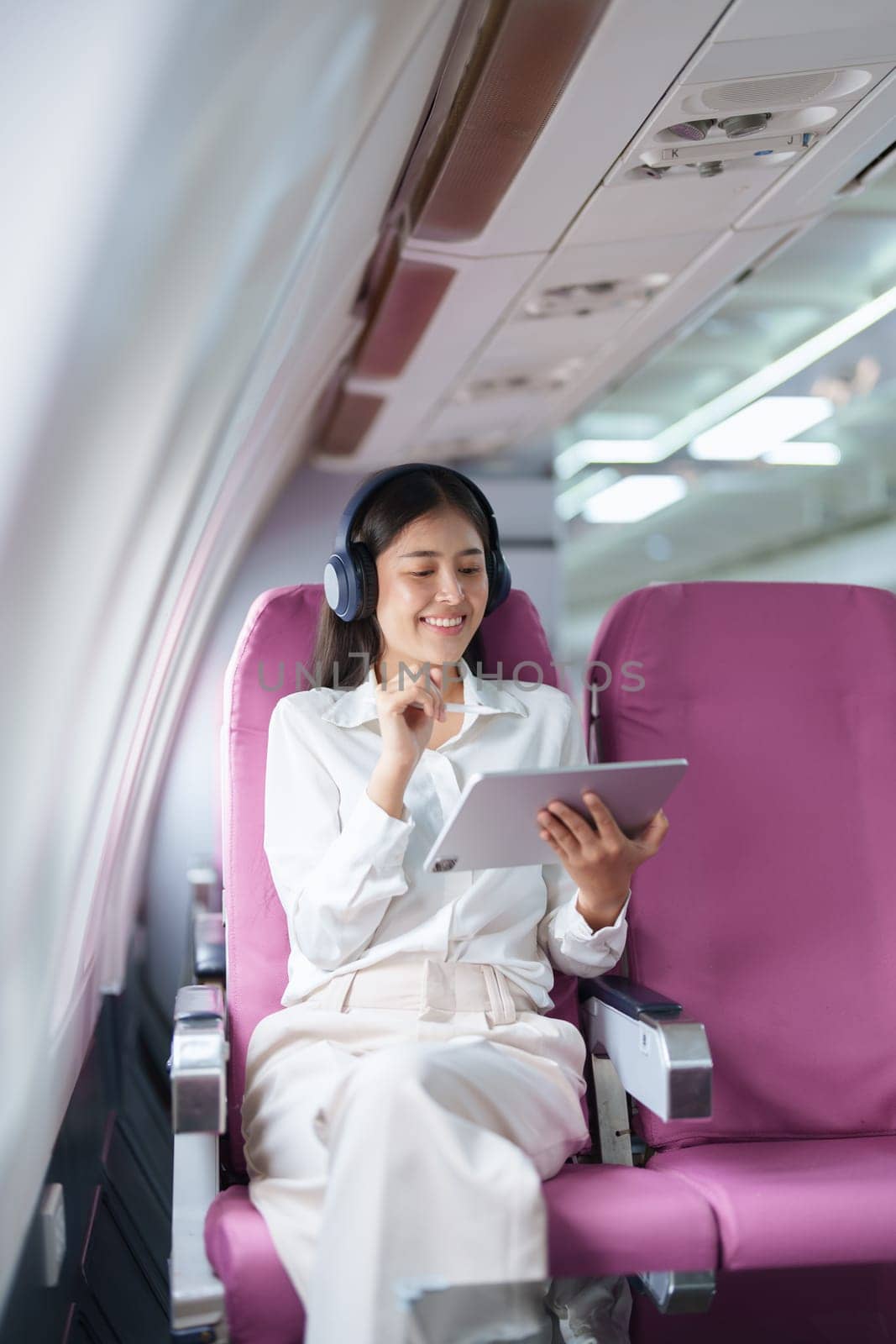 Young Asian attractive business woman passenger sitting on business class luxury plane while wearing wireless headphone for relax and working with tablet during the flight by Manastrong