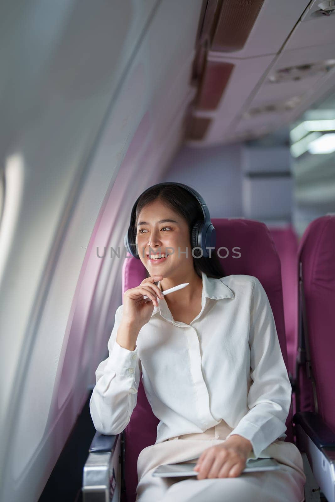 Young Asian attractive business woman passenger sitting on business class luxury plane while wearing wireless headphone for relax and working with tablet during the flight.