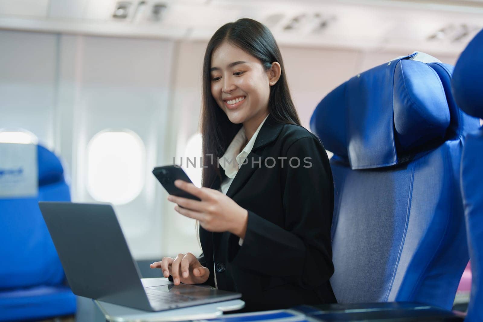 Asian business woman passenger sitting on business class luxury plane while working using smartphone mobile talking and use laptop computer with simulated space using on board wireless connection by Manastrong