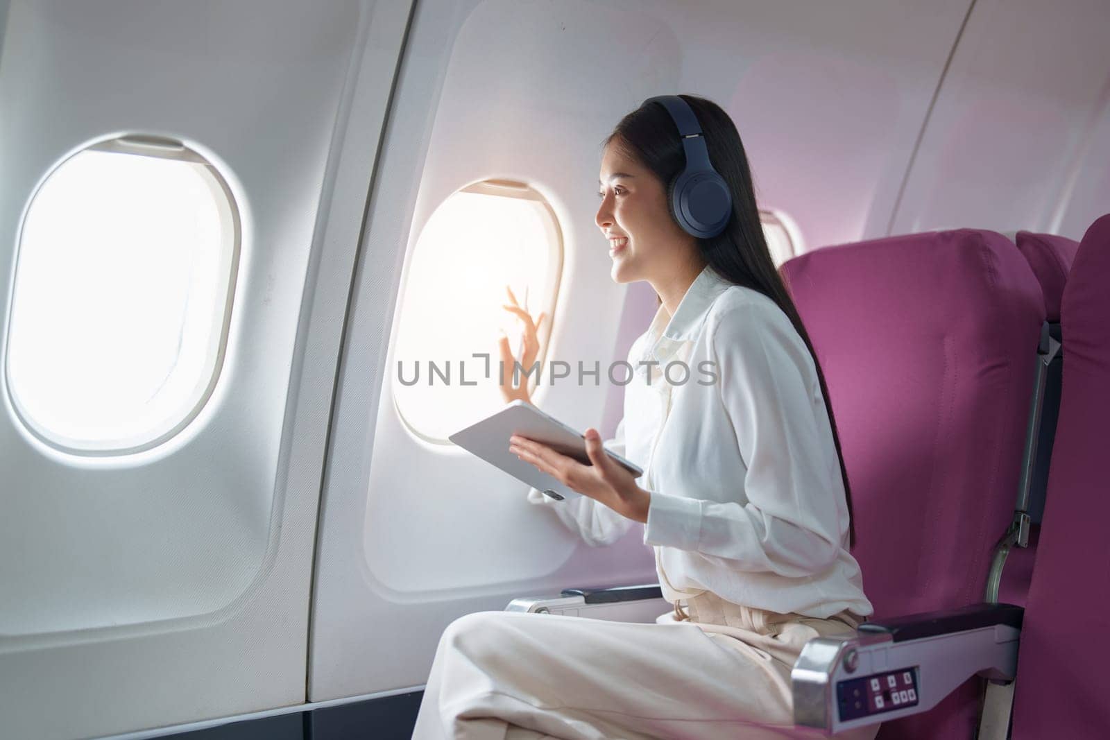 Young Asian attractive business woman passenger sitting on business class luxury plane while wearing wireless headphone for relax and working with tablet during the flight by Manastrong