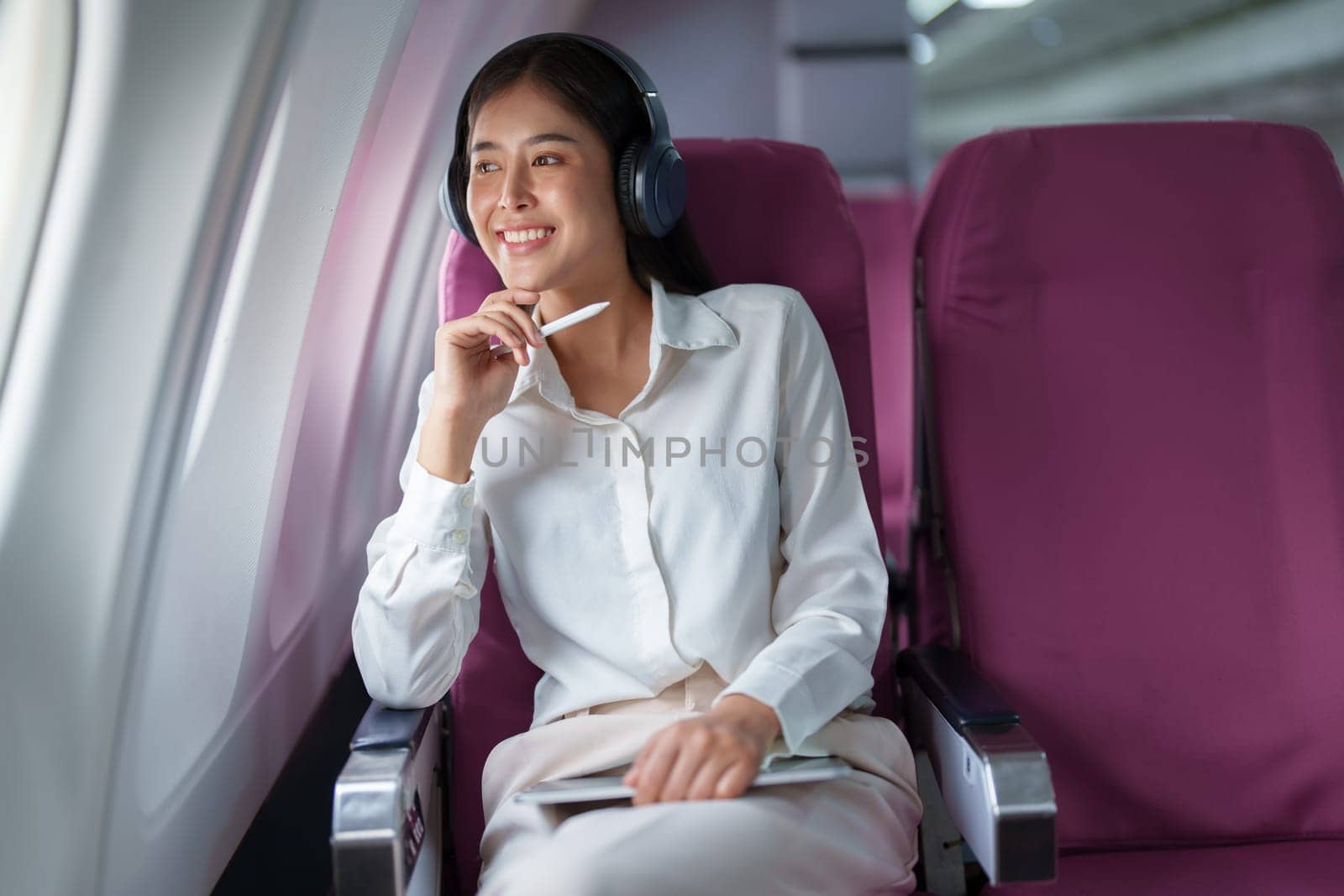 Young Asian attractive business woman passenger sitting on business class luxury plane while wearing wireless headphone for relax and working with tablet during the flight by Manastrong
