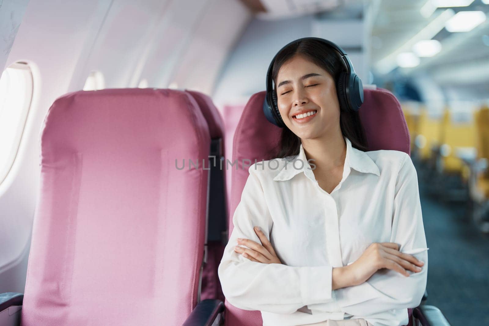 Young Asian attractive business woman passenger sitting on business class luxury plane while wearing wireless headphone for relax and working with tablet during the flight.