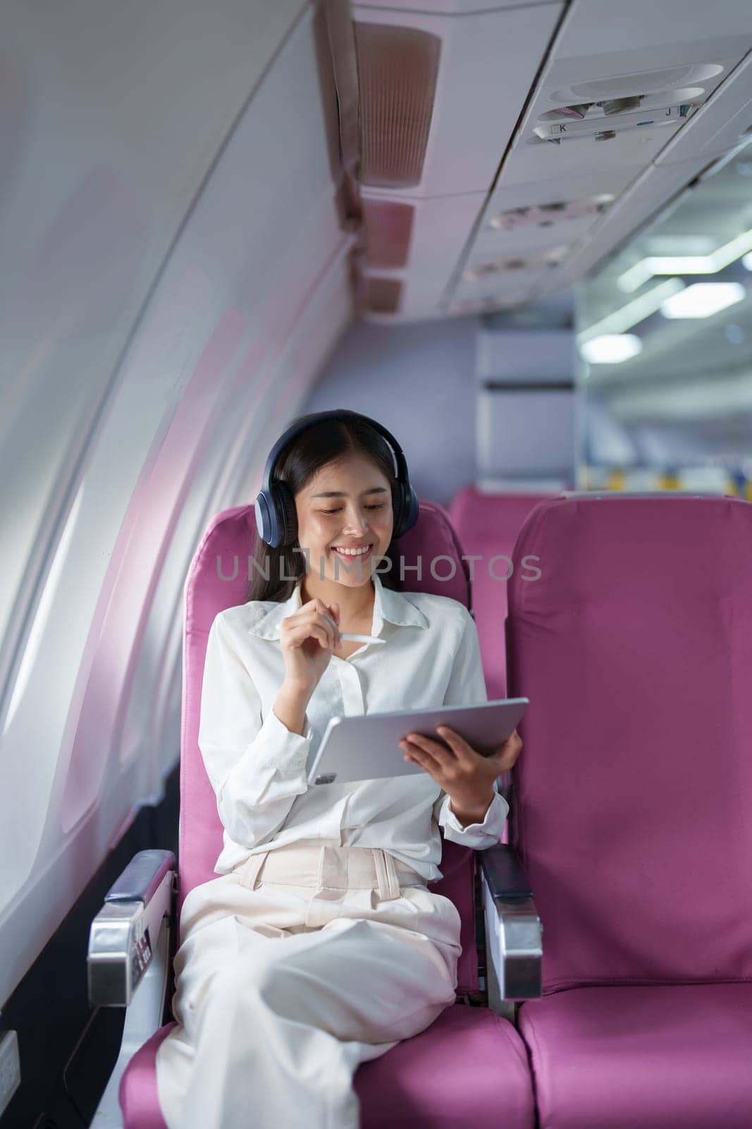 Young Asian attractive business woman passenger sitting on business class luxury plane while wearing wireless headphone for relax and working with tablet during the flight.