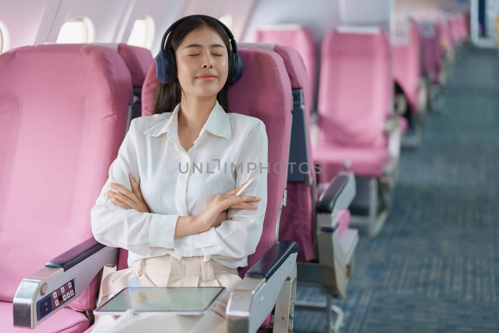 Young Asian attractive business woman passenger sitting on business class luxury plane while wearing wireless headphone for relax and working with tablet during the flight.