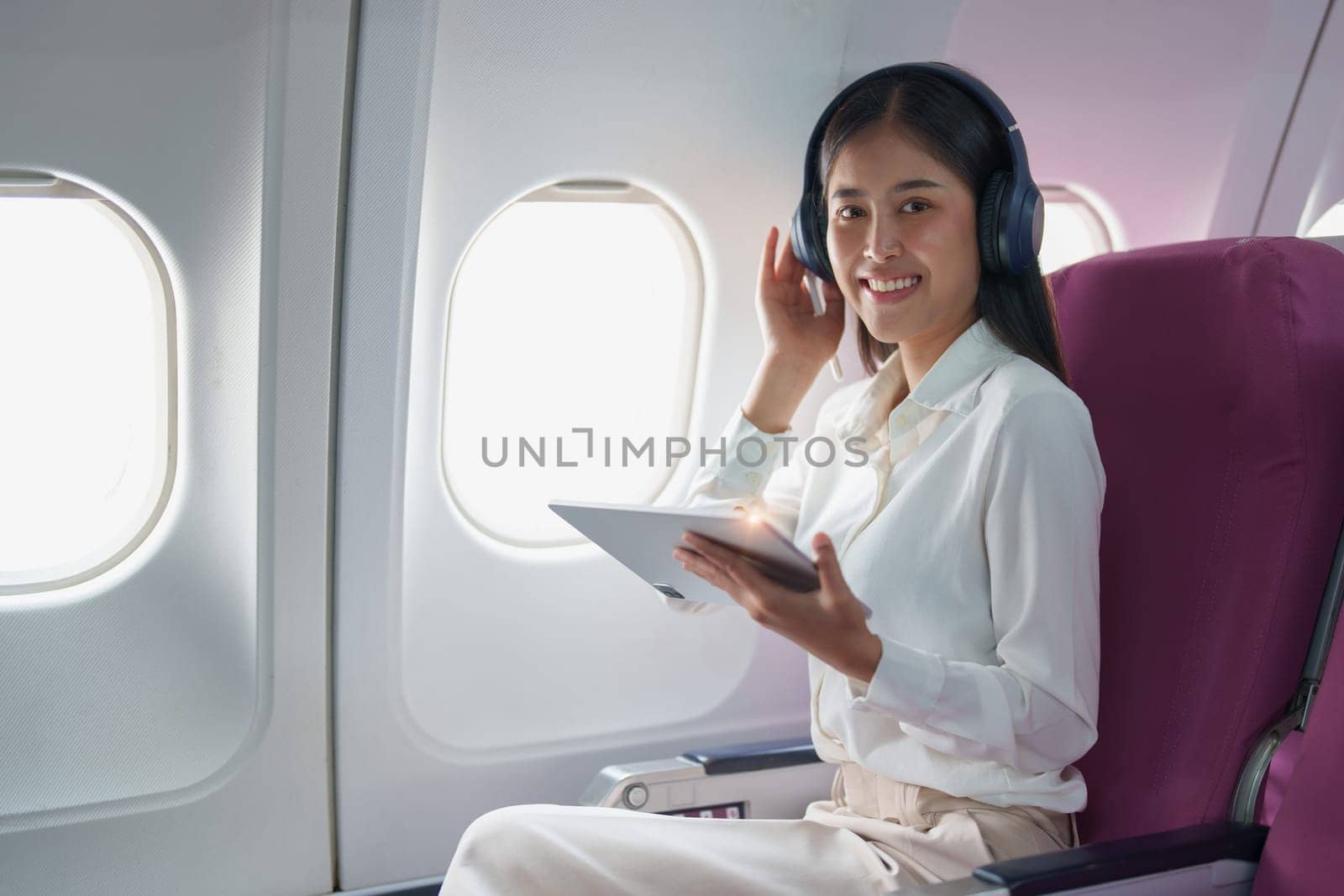 Young Asian attractive business woman passenger sitting on business class luxury plane while wearing wireless headphone for relax and working with tablet during the flight by Manastrong