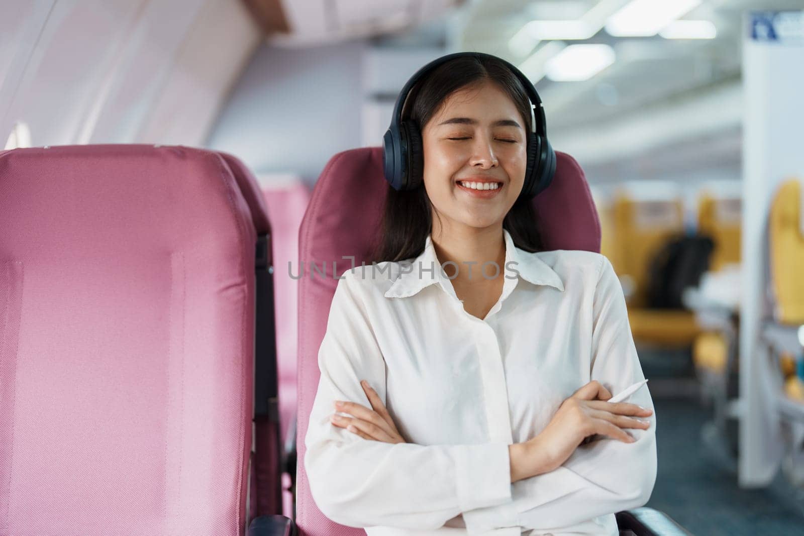 Young Asian attractive business woman passenger sitting on business class luxury plane while wearing wireless headphone for relax and working with tablet during the flight.