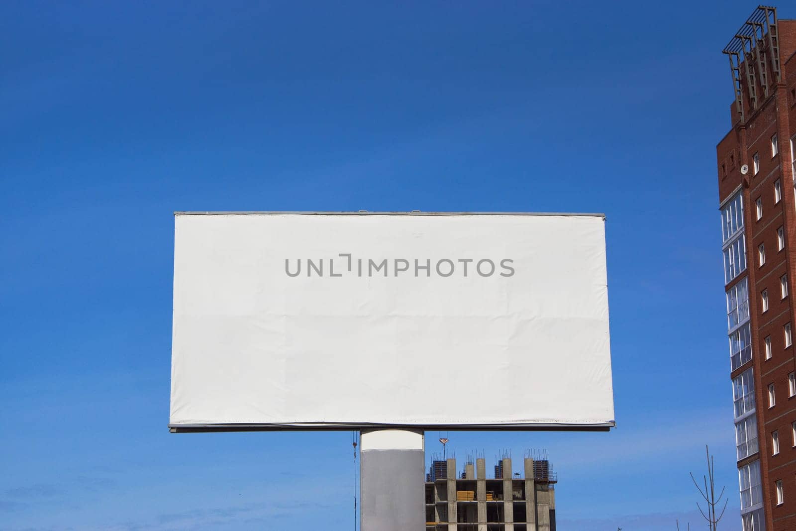 A large empty mock-up of a billboard next to buildings and residential buildings under construction against a blue sky background.