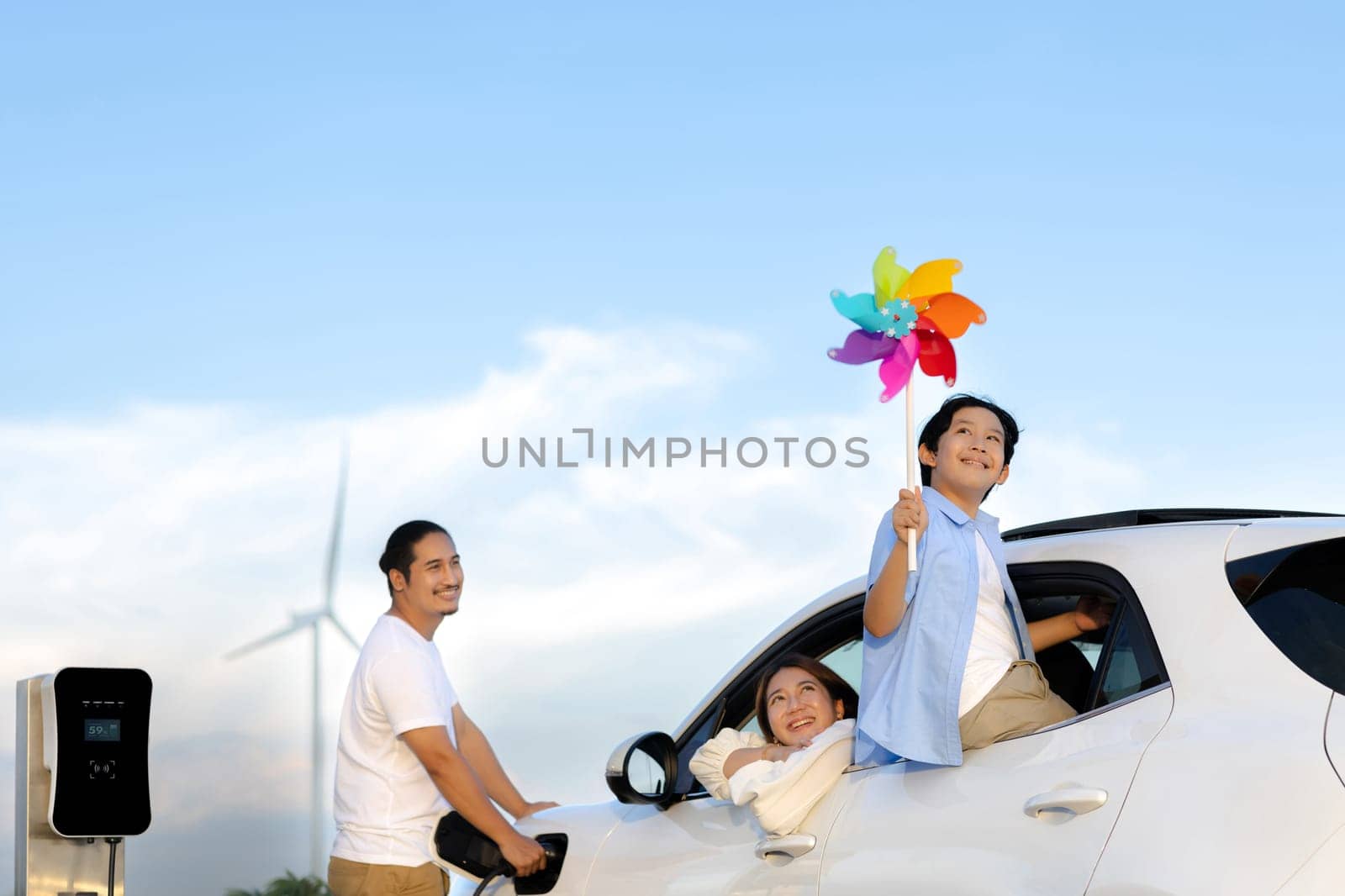 Concept of progressive happy family enjoying their time at wind farm with electric vehicle. Electric vehicle driven by clean renewable energy from wind turbine generator for charging station.