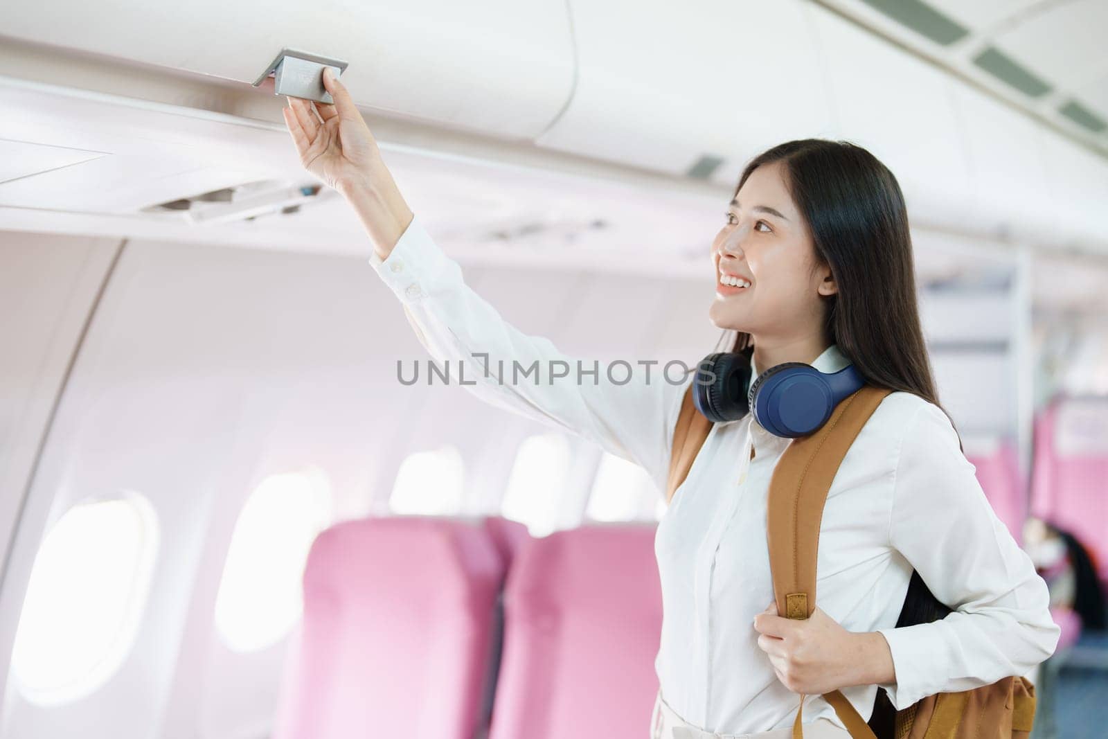 Young asian attractive woman travel by airplane, Passenger wearing headphone putting hand baggage in lockers above seats of plane.