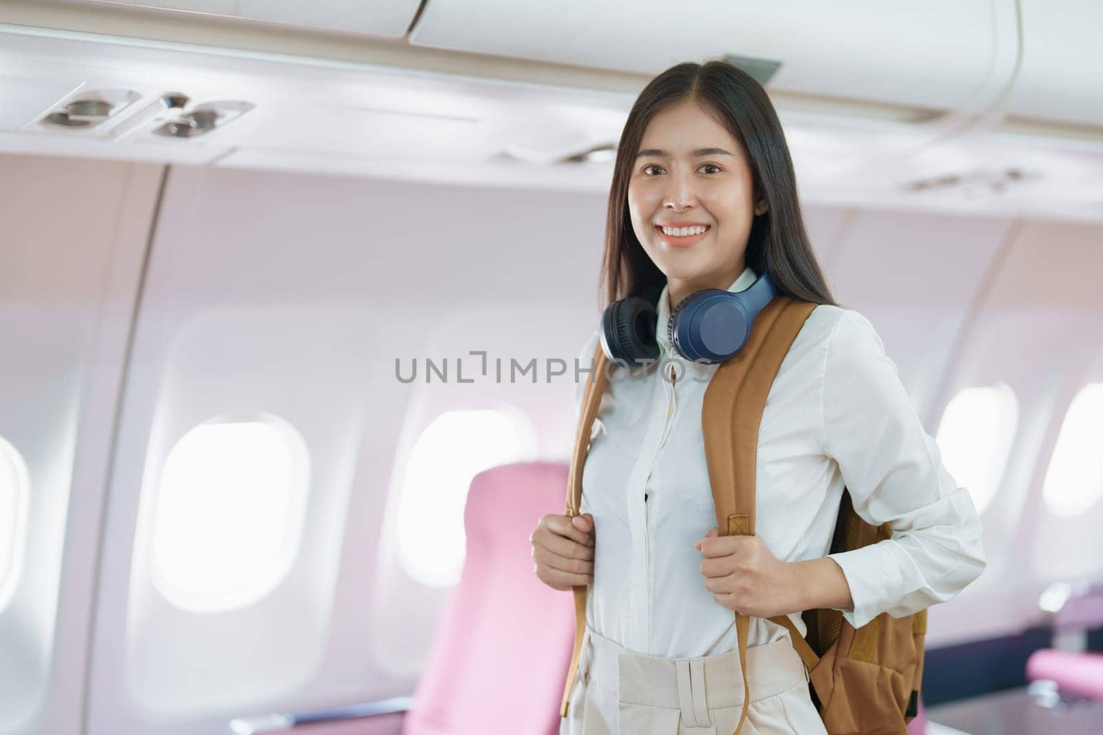 Young asian attractive woman travel by airplane, Passenger wearing headphone putting hand baggage in lockers above seats of plane.