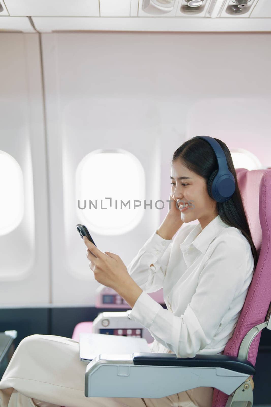 Young Asian attractive business woman passenger sitting on business class luxury plane while wearing wireless headphone and using smart phone mobile for relax during the flight.