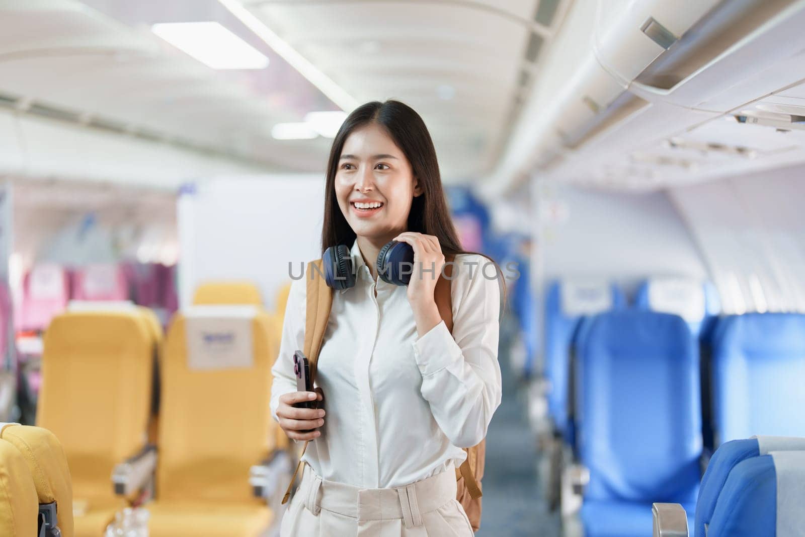 Young asian attractive woman travel by airplane, Passenger wearing headphone putting hand baggage in lockers above seats of plane.