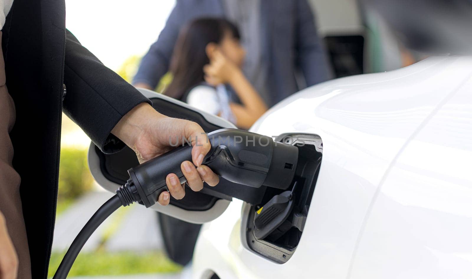 Focus closeup hand charging electric car, insert charger device into electric vehicle as progressive lifestyle concept of alternative green energy technology with blurred family in the background.