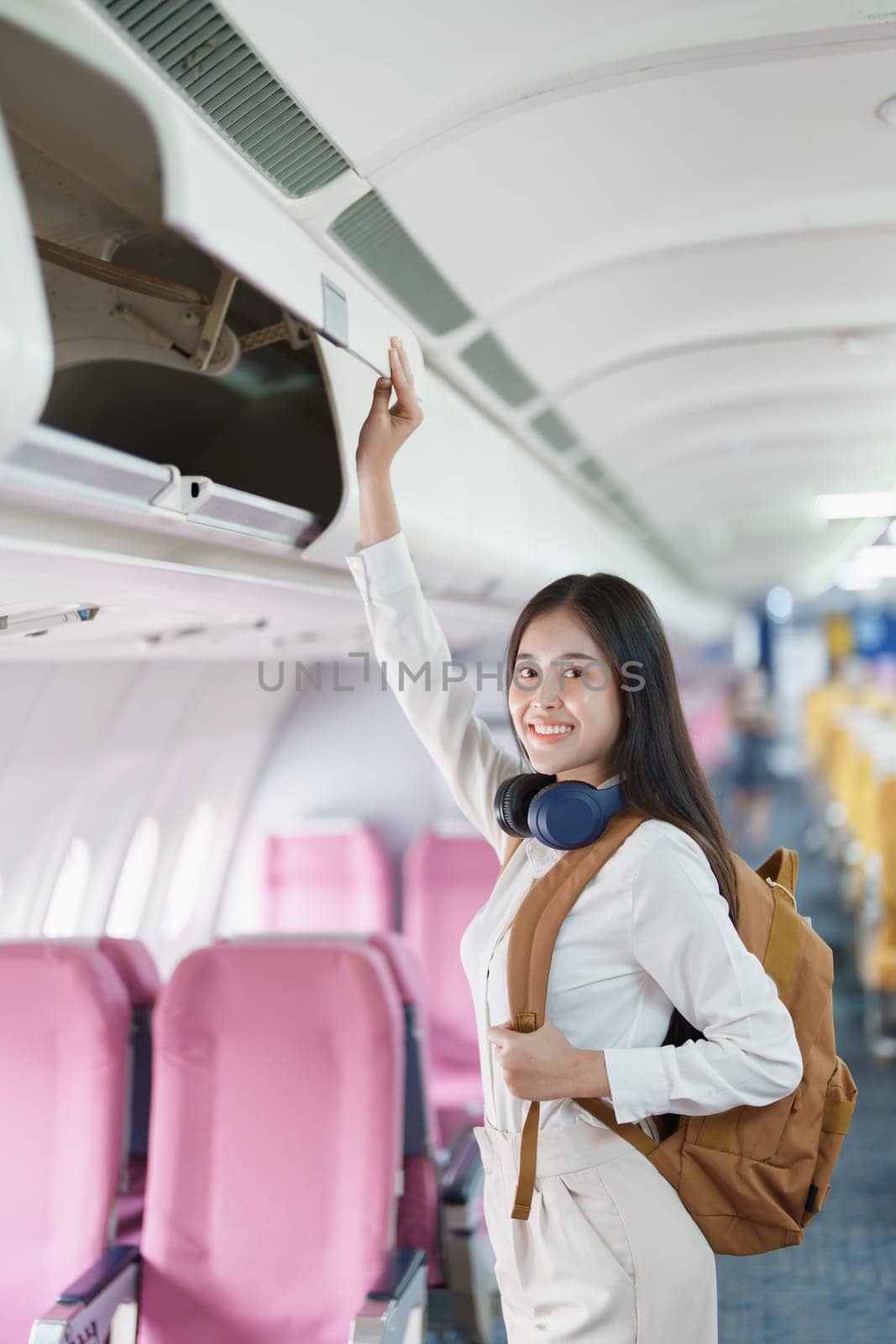 Young asian attractive woman travel by airplane, Passenger wearing headphone putting hand baggage in lockers above seats of plane by Manastrong