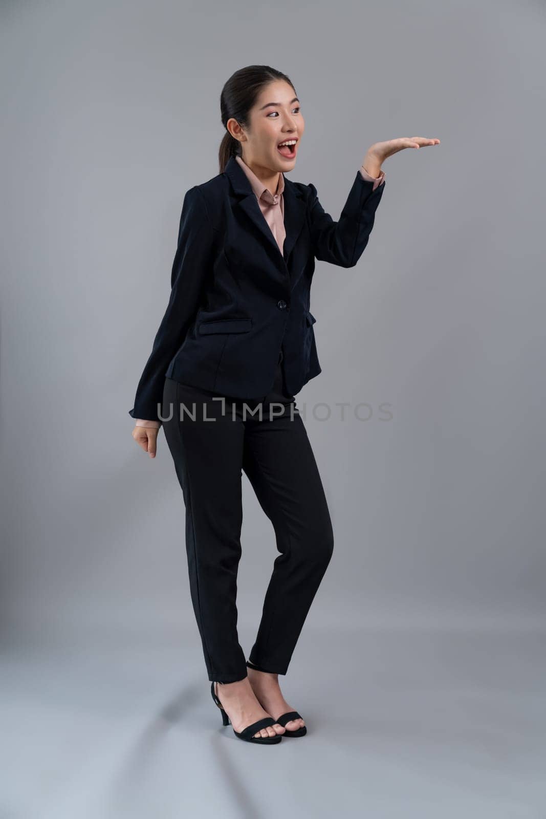 Confident young asian businesswoman in formal suit holding hand to indicate promotion or advertising with amazed expression and gesture on isolated background. Enthusiastic