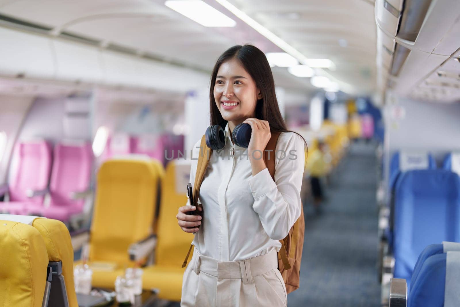 Young asian attractive woman travel by airplane, Passenger wearing headphone putting hand baggage in lockers above seats of plane.