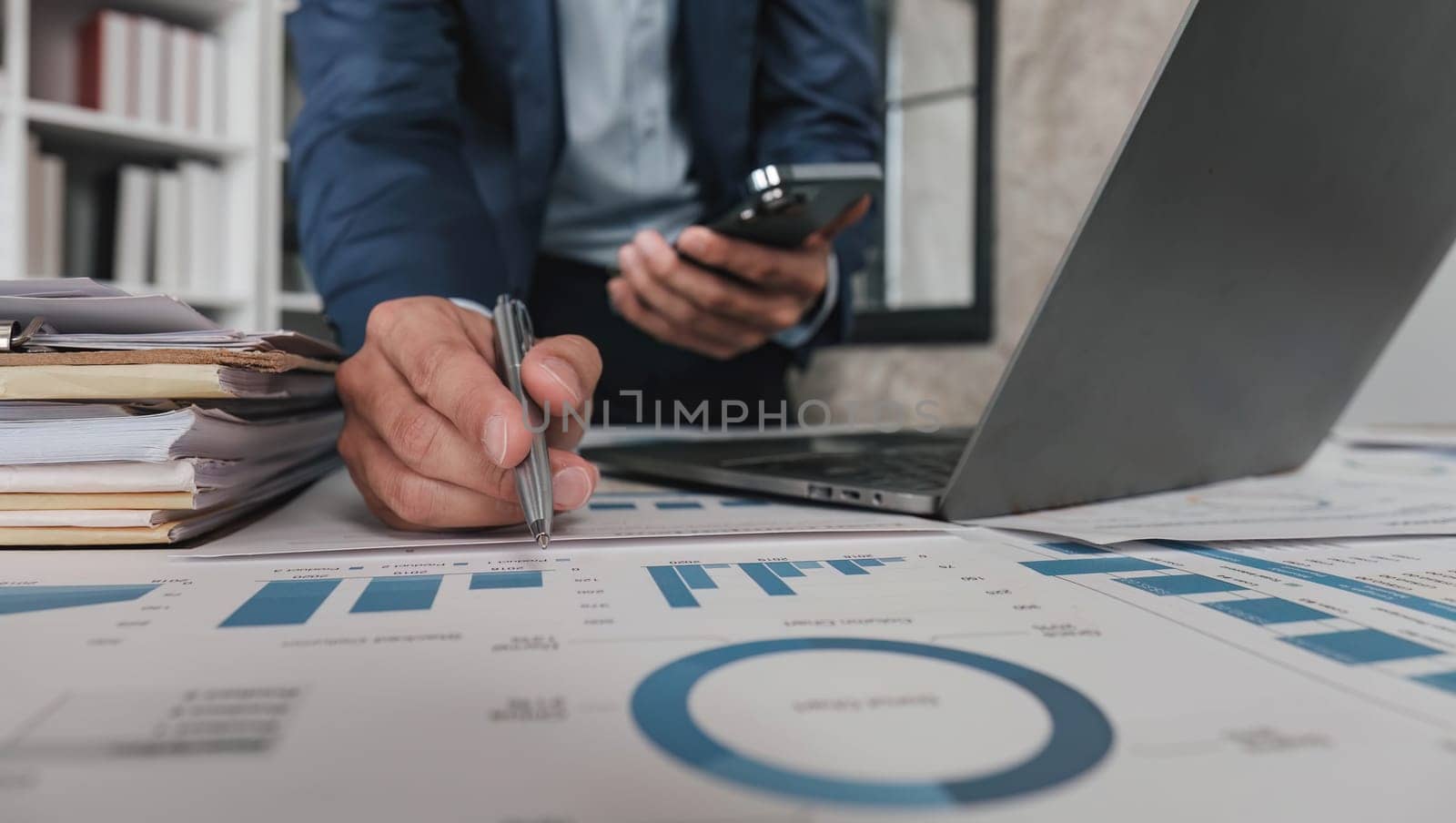 Businessman pointing pen at data graph and use moblie phone, estimating profit and income statement of business in office...