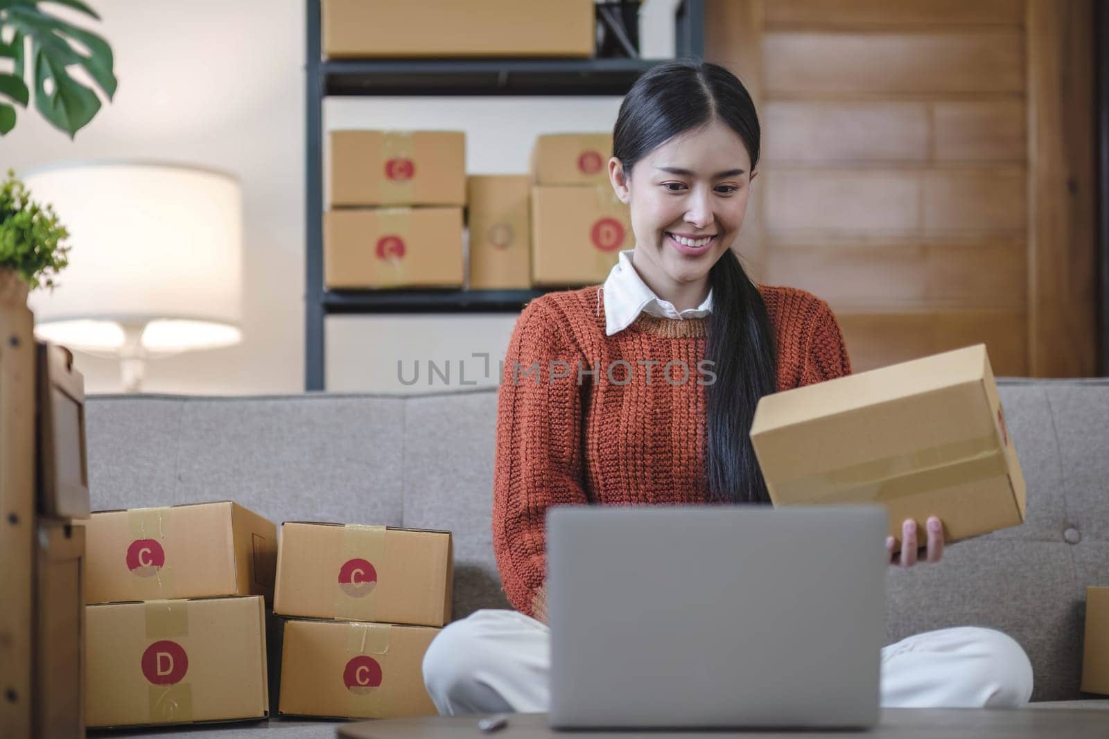 Asian business woman leaning on the sofa use laptop computer checking customer order online shipping boxes at home. Starting Small business entrepreneur SME. Online business, Work at home concept. by wichayada