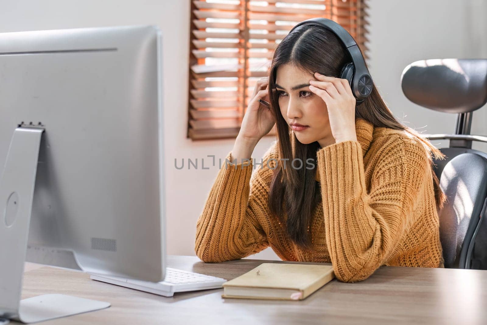 Tired teen asian lady in headphones falls asleep at table with laptop in room interior. Boring video lesson at home, problems at study, education with modern technology, online webinar and overwork by wichayada