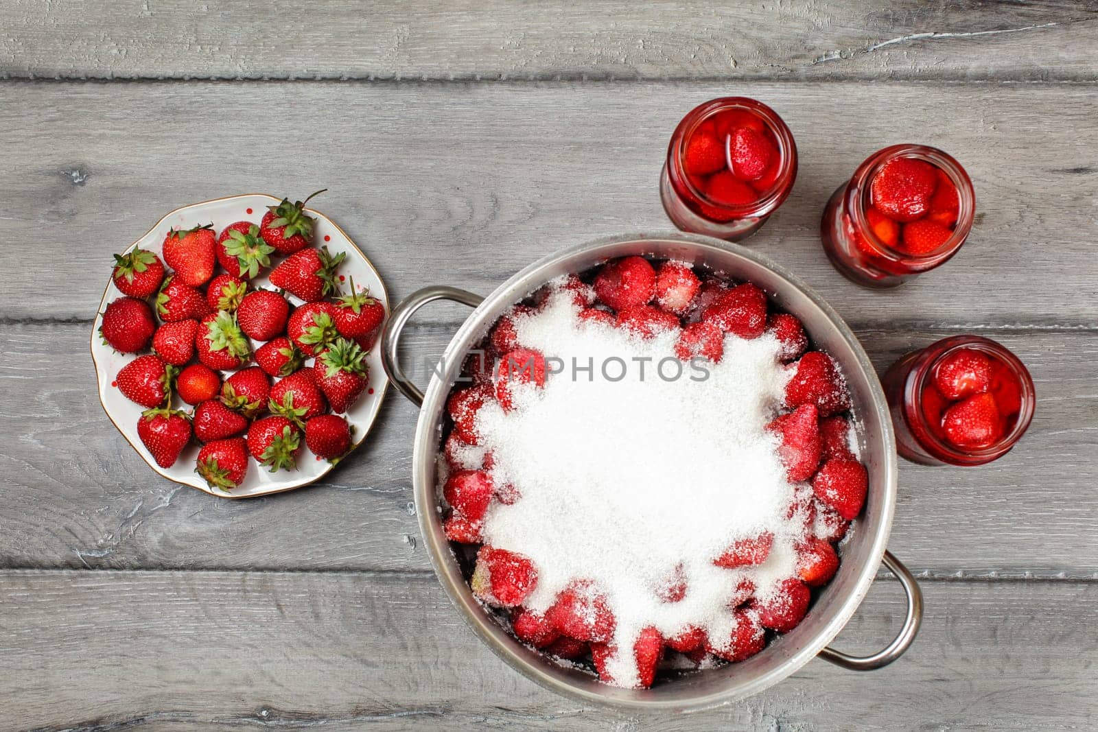 Tabletop view, large steel pot with strawberries covered in crystal sugar, three flasks with fruit in syrup and plate next to it. Homemade strawberry compote preparation.