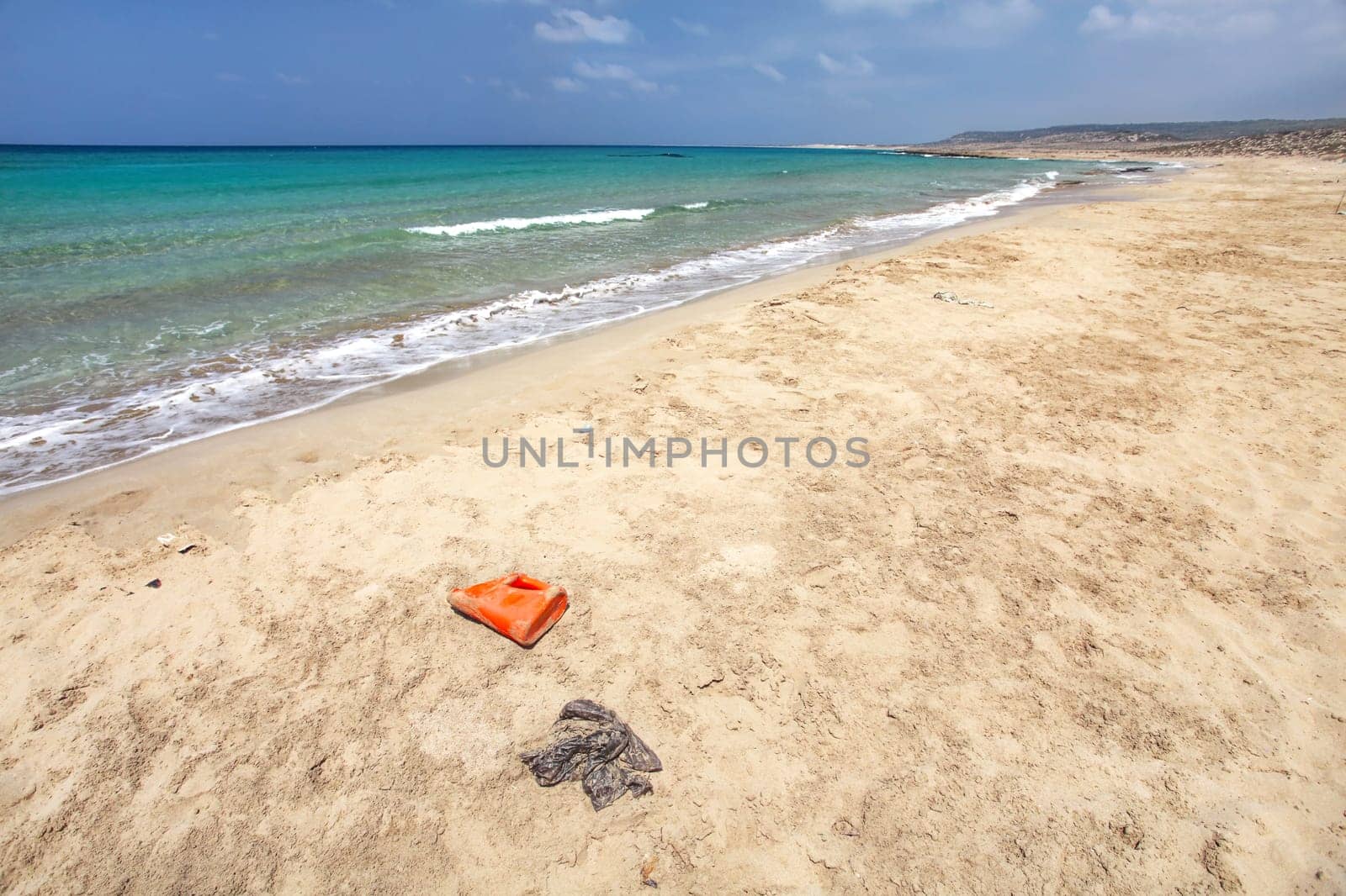 Beautiful wild beach with no tourists, crystal clear sea, but waste (plastic red bottle, black bag and other small debris) spoils the perfect picture. Ocean pollution concept.