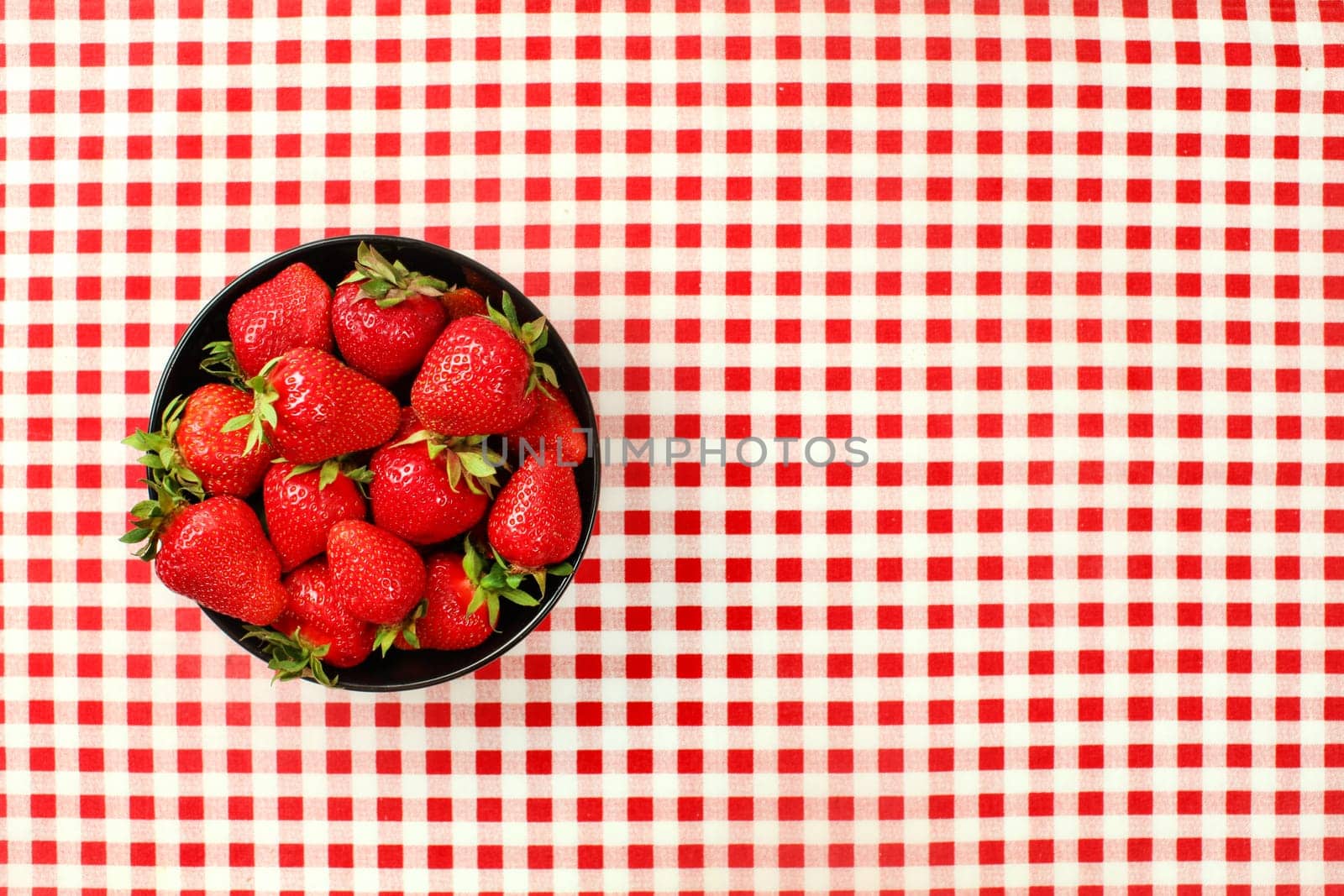 Tabletop view, small black ceramic bowl of strawberries on red chequered gingham tablecloth