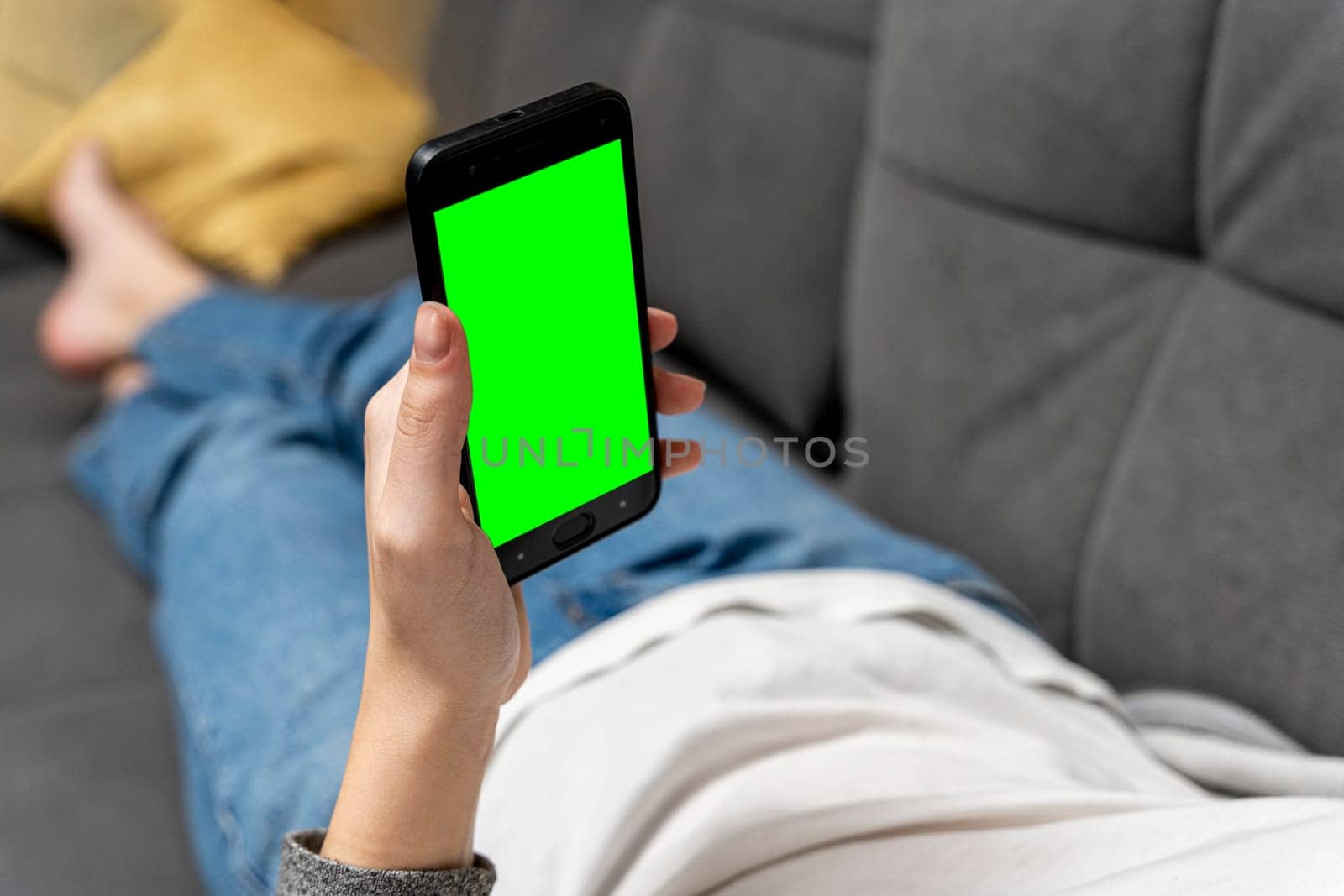 an unrecognizable girl uses a smartphone with a green screen while lying at home on the couch. a smartphone with a chromakey in female hands. View videos and photos on social networks. web browsing on the Internet, browsing. Dependence on gadgets