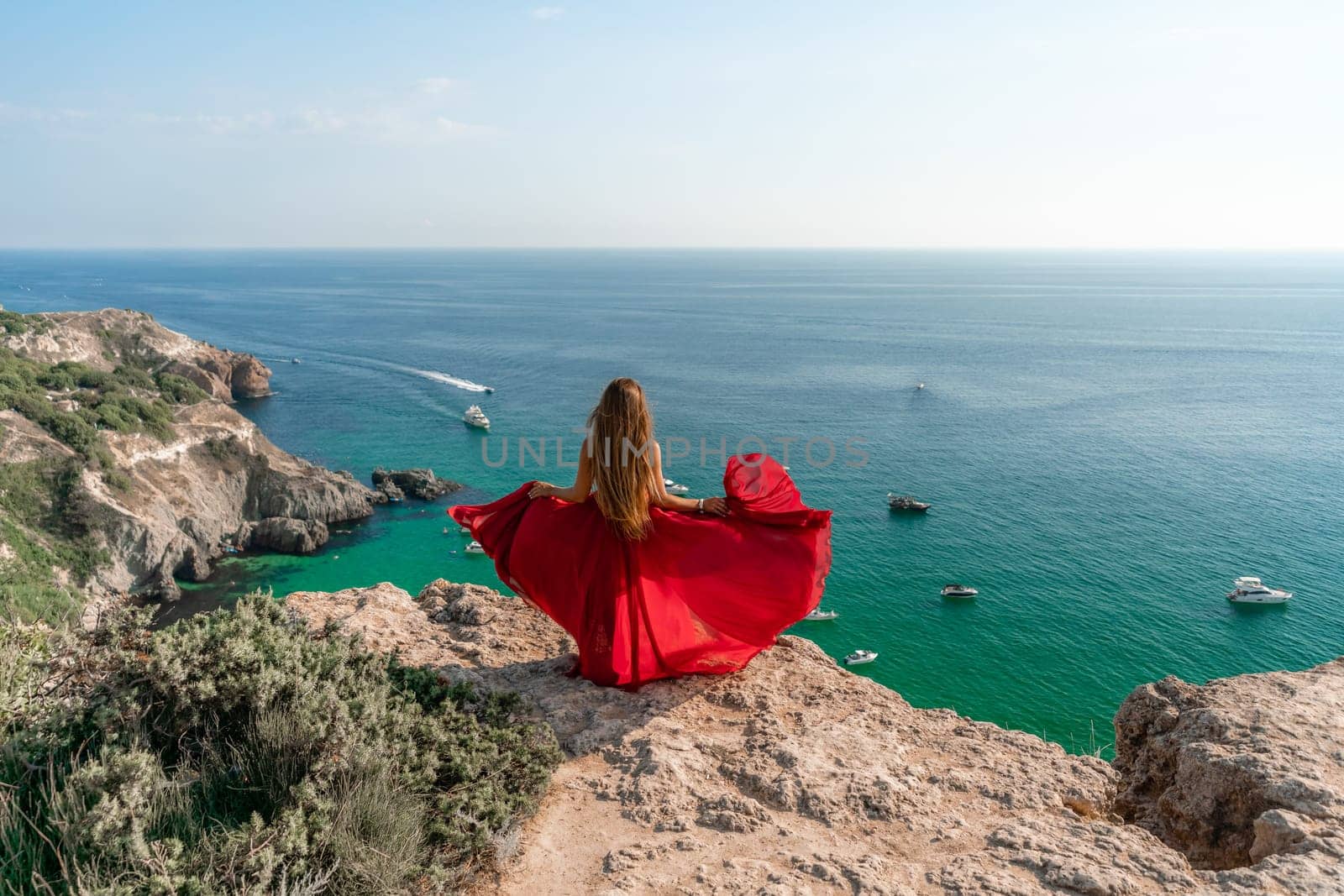 Woman in a red dress on the sea. Rear view of a happy woman in a red long dress posing on a rock high above the sea sees a yacht. Girl in nature against the blue sky. by Matiunina