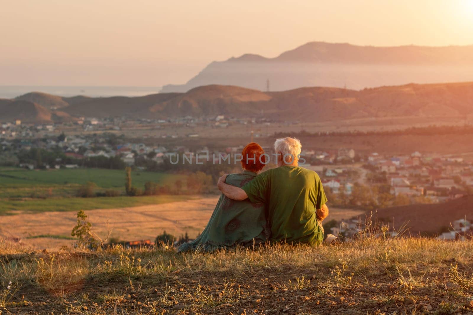 An elderly couple sits on a mountain with their backs with a beautiful view of the mountains and the sea in the distance