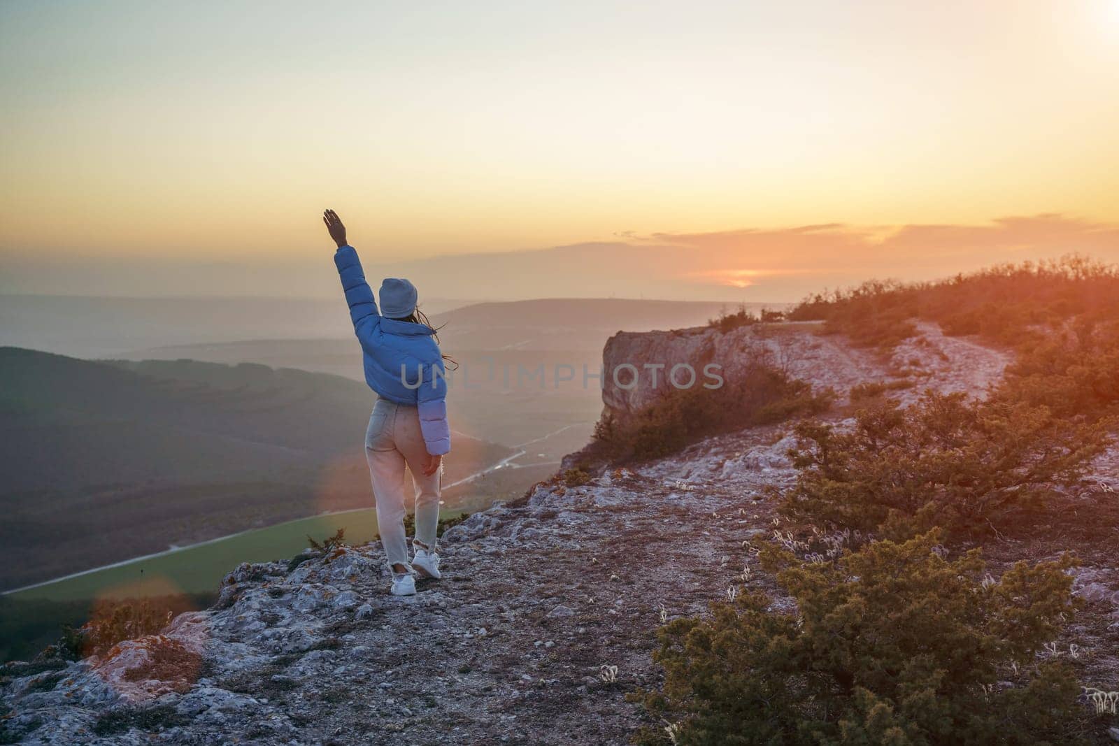 Woman hiker open arms on top of sunrise mountain. The girl salutes the sun, wearing a blue jacket, white hat and white jeans. Conceptual design
