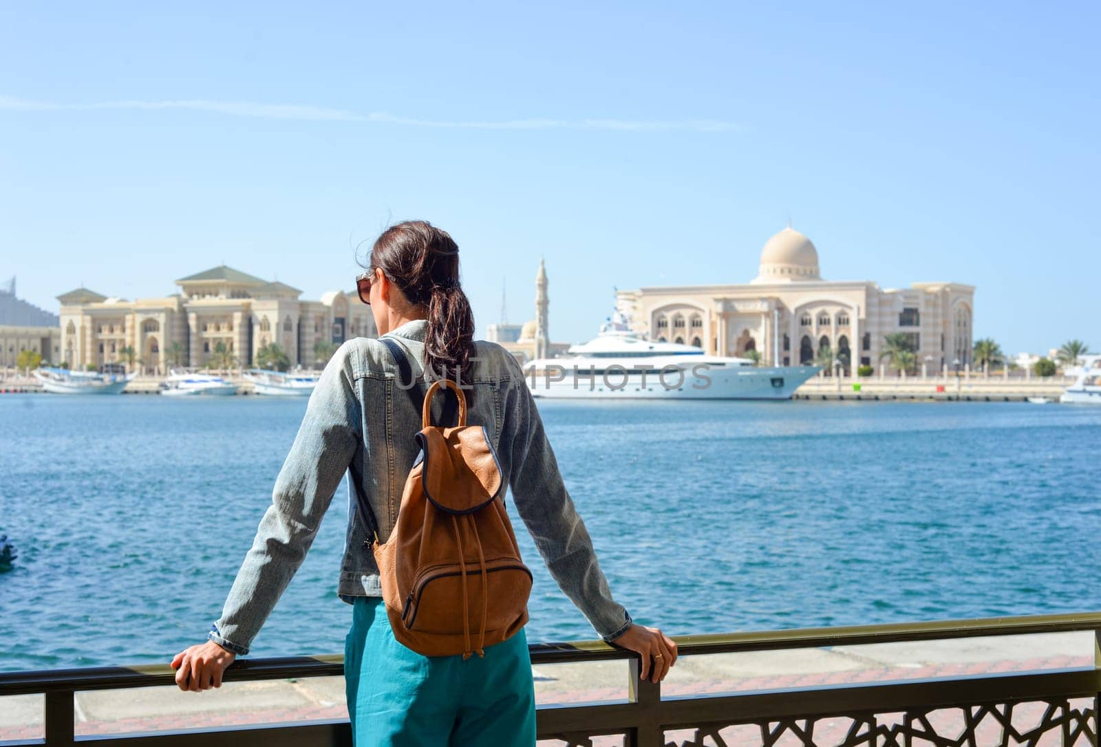 A young woman tourist with a backpack looks at the administrative region of the emirate of Sharjah with the port and ships