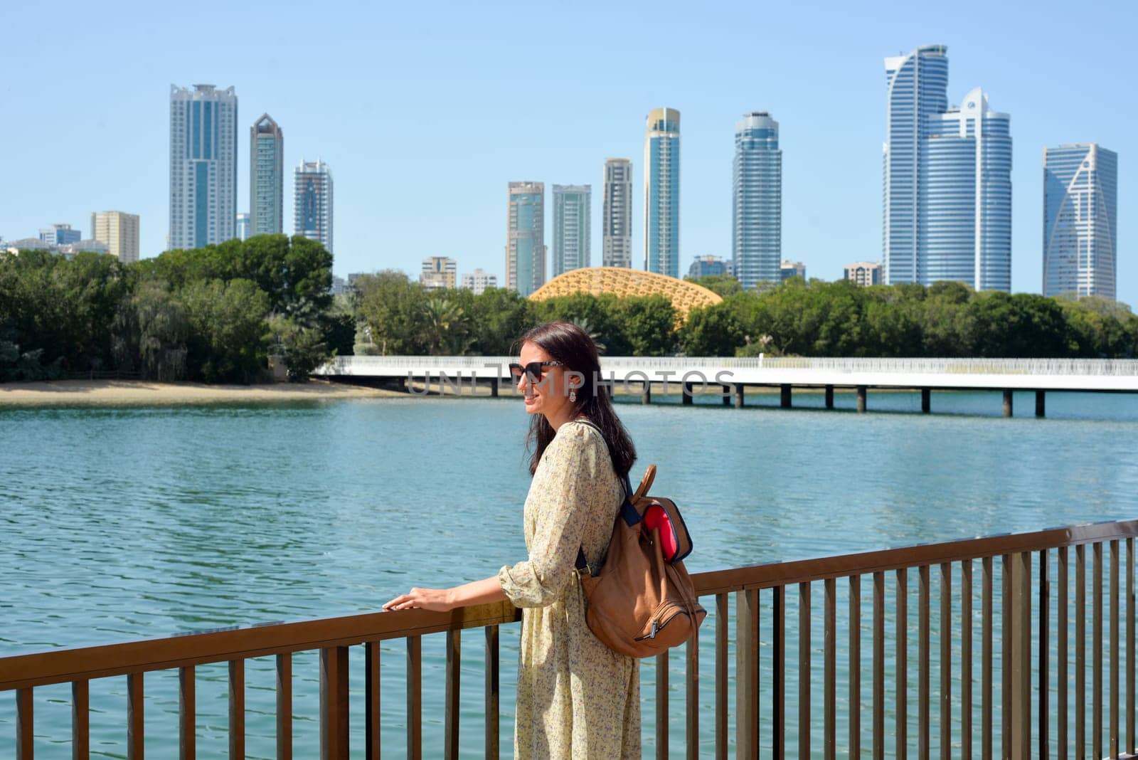 The skyline of Sharjah is visible from the waterfront - a woman tourist with a backpack enjoys the view of the water and skyscrapers. Cityscape of Sharjah UAE