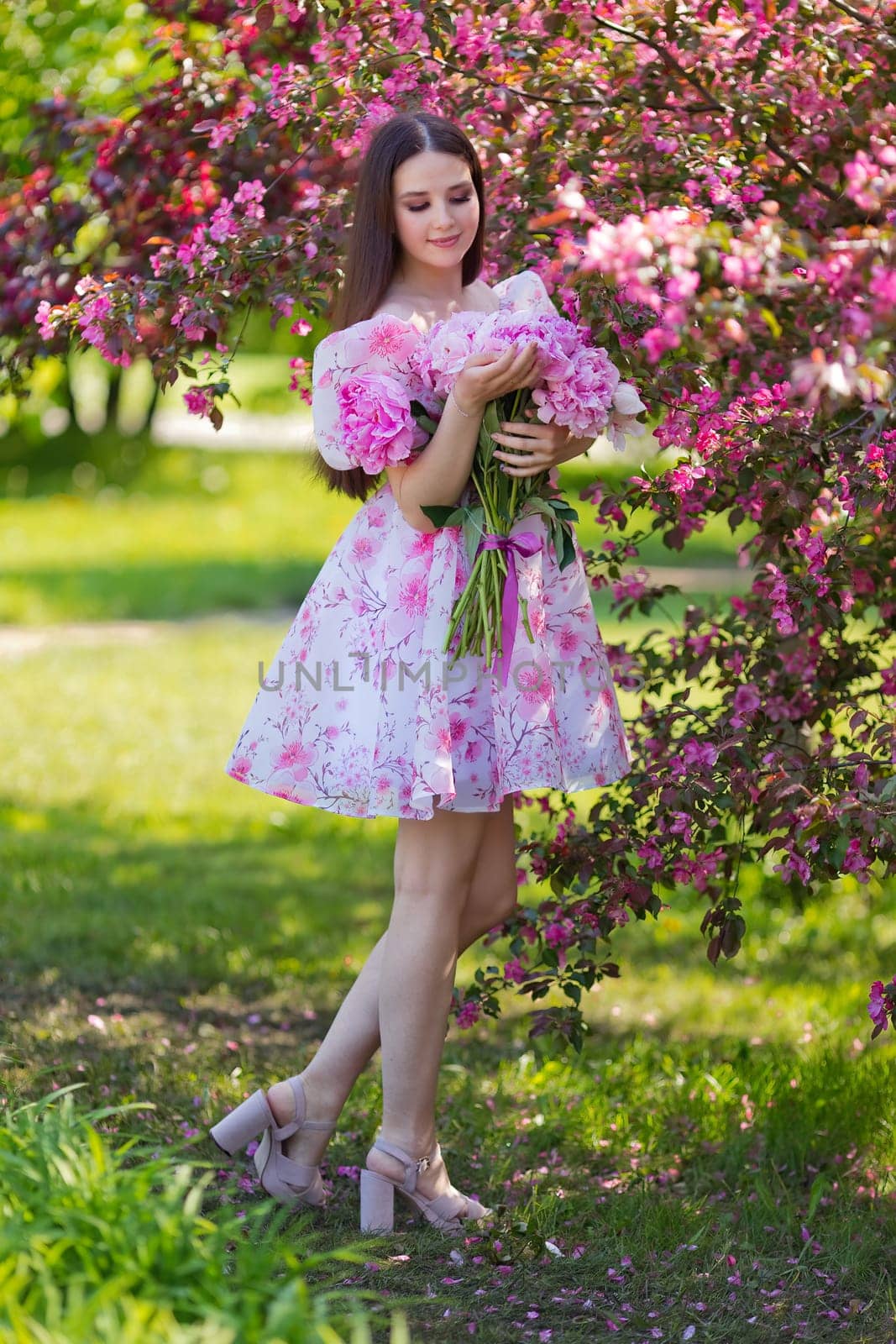 A beautiful brunette in a light pink dress, with a large bouquet of pink peonies, stands near pink blooming trees, in the garden on a sunny day. Copy space. Vertical