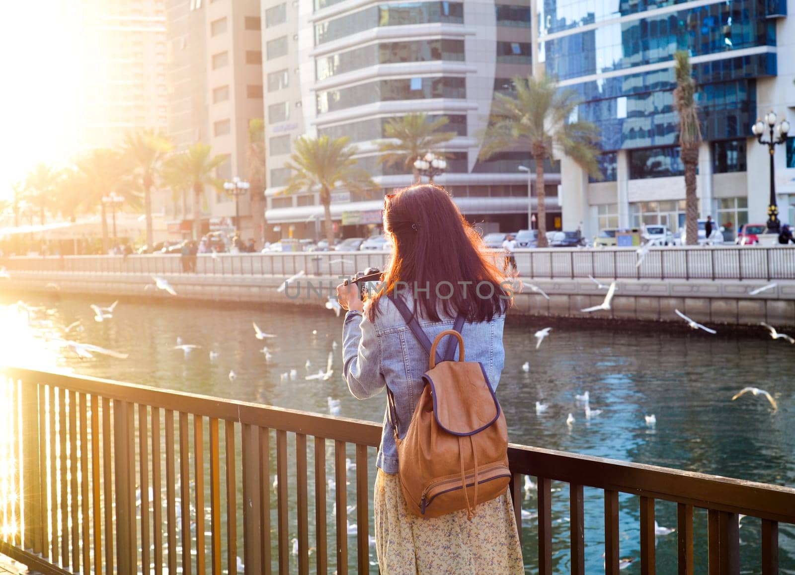 A woman in a long dress with a backpack photographs seagulls on the Al Kasbah Canal in the emirate of Sharjah at sunset. Back view