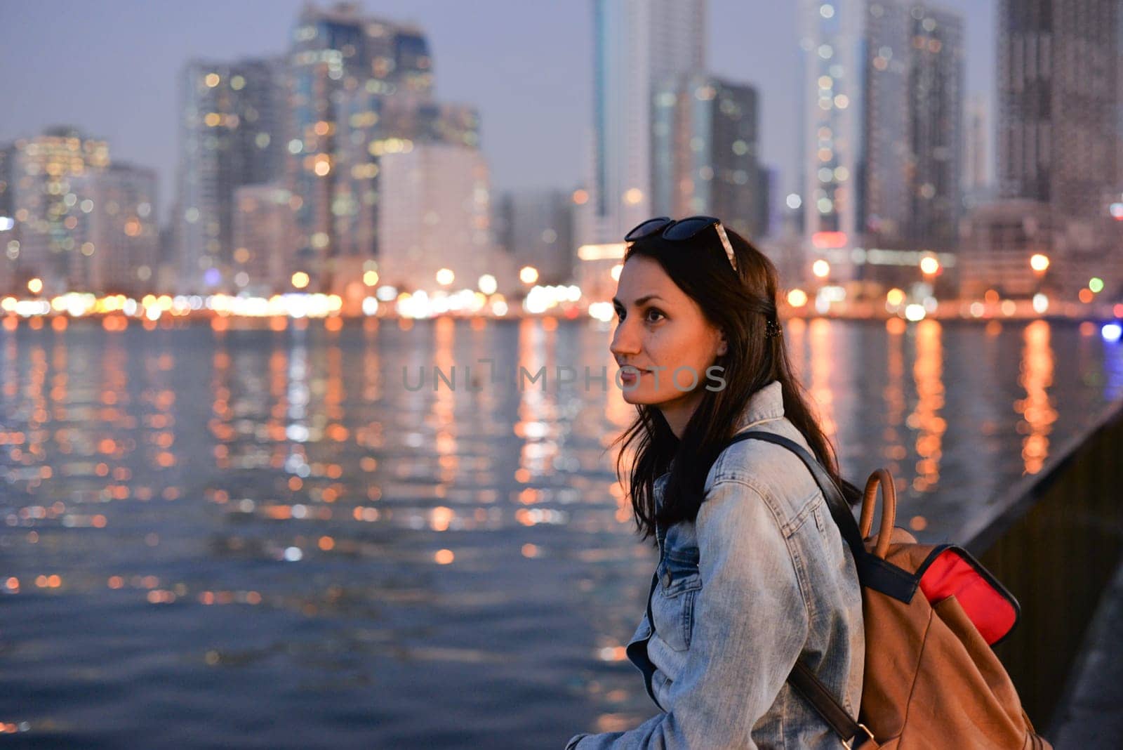 A tourist girl with a backpack on her shoulders enjoys a view of the modern skyscrapers of the Sharjah marina at night.