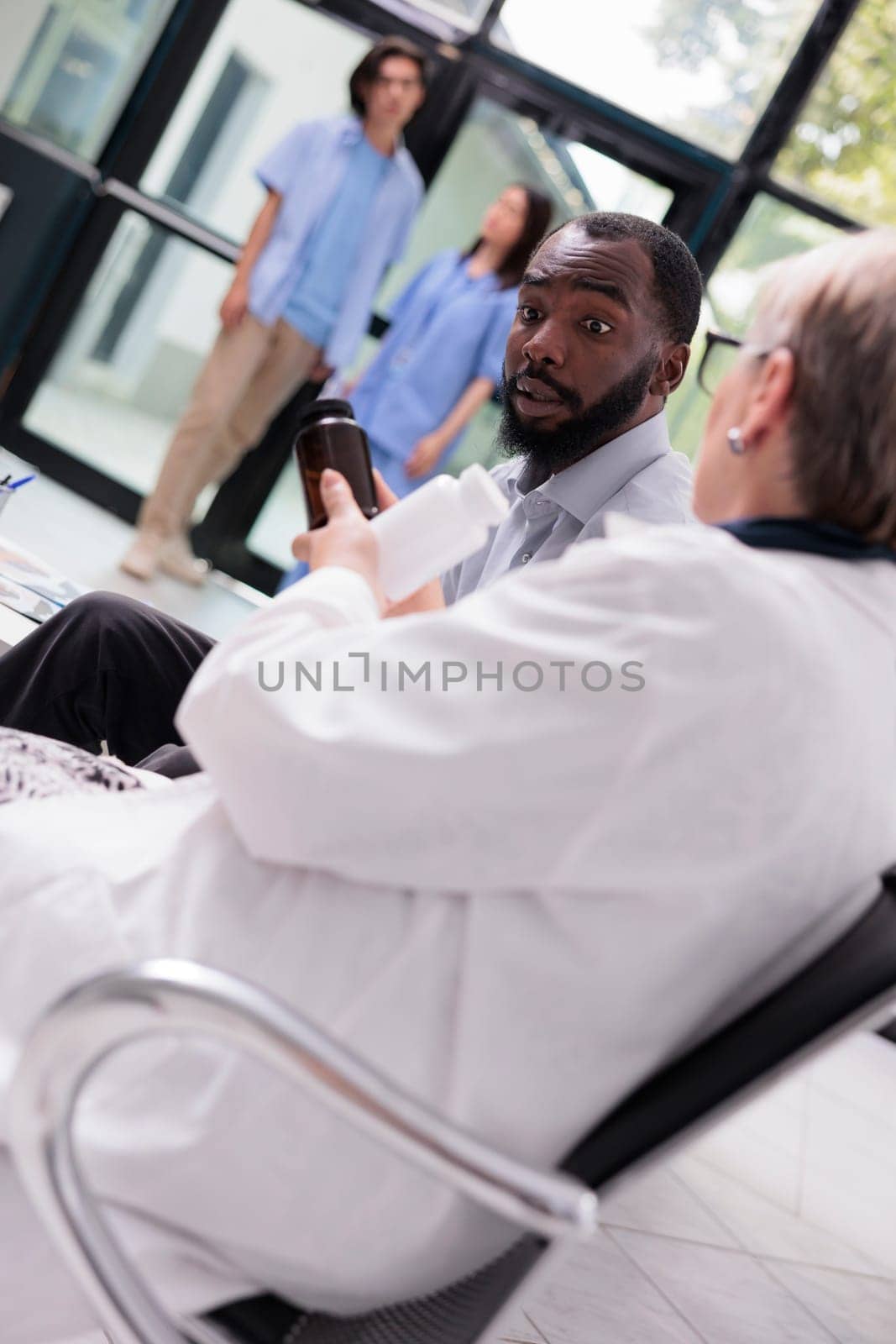 Senior specialist doctor discussing health care treatment with patient while showing pills bottle during consultation in hospital waiting area. Diverse people standing in lobby, medicine service
