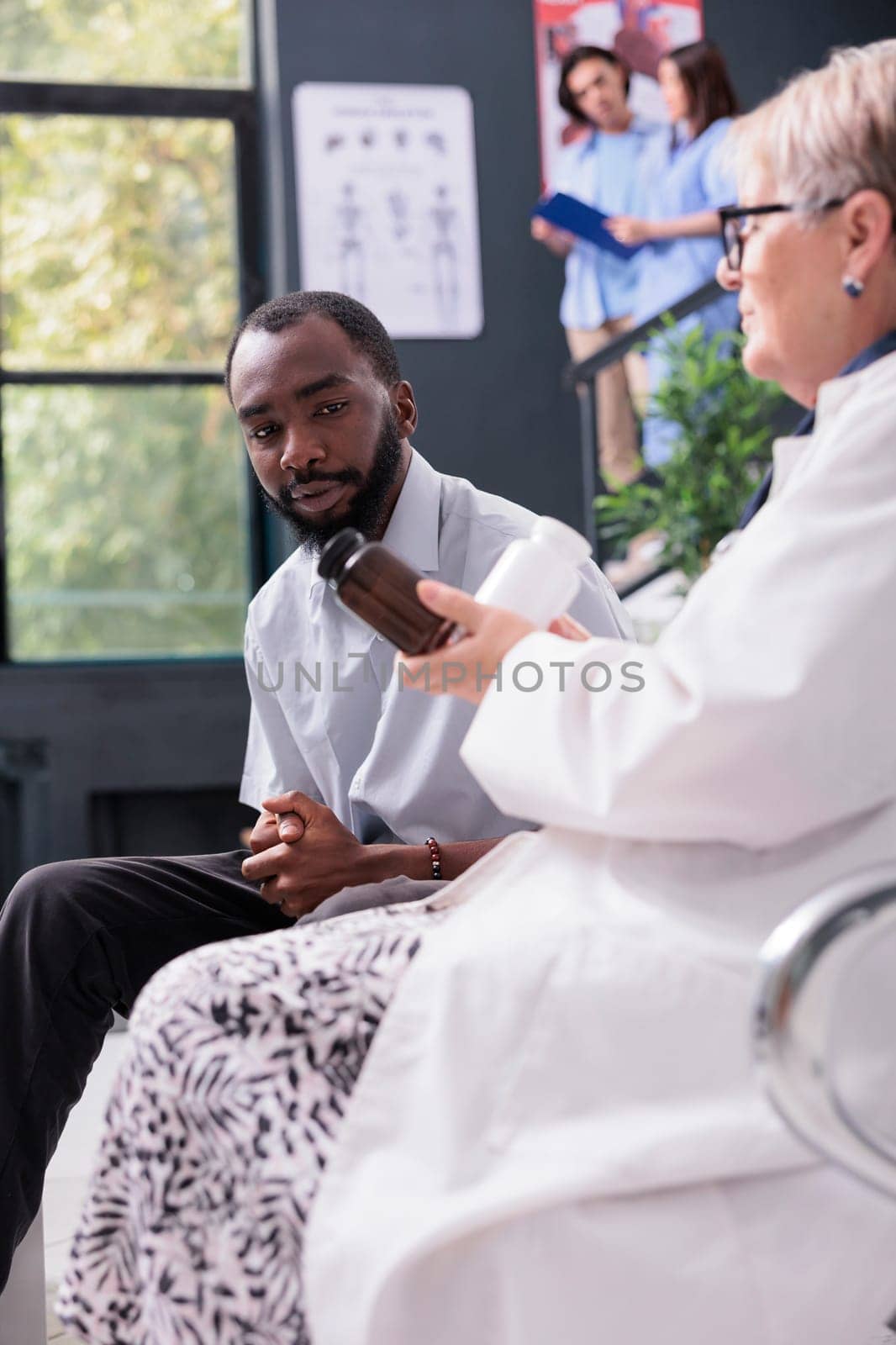 African american patient sitting on chair in hospital waiting area while medic explaining medication treatment during examination. Senior physician doctor showing pill bottles full with painkiller