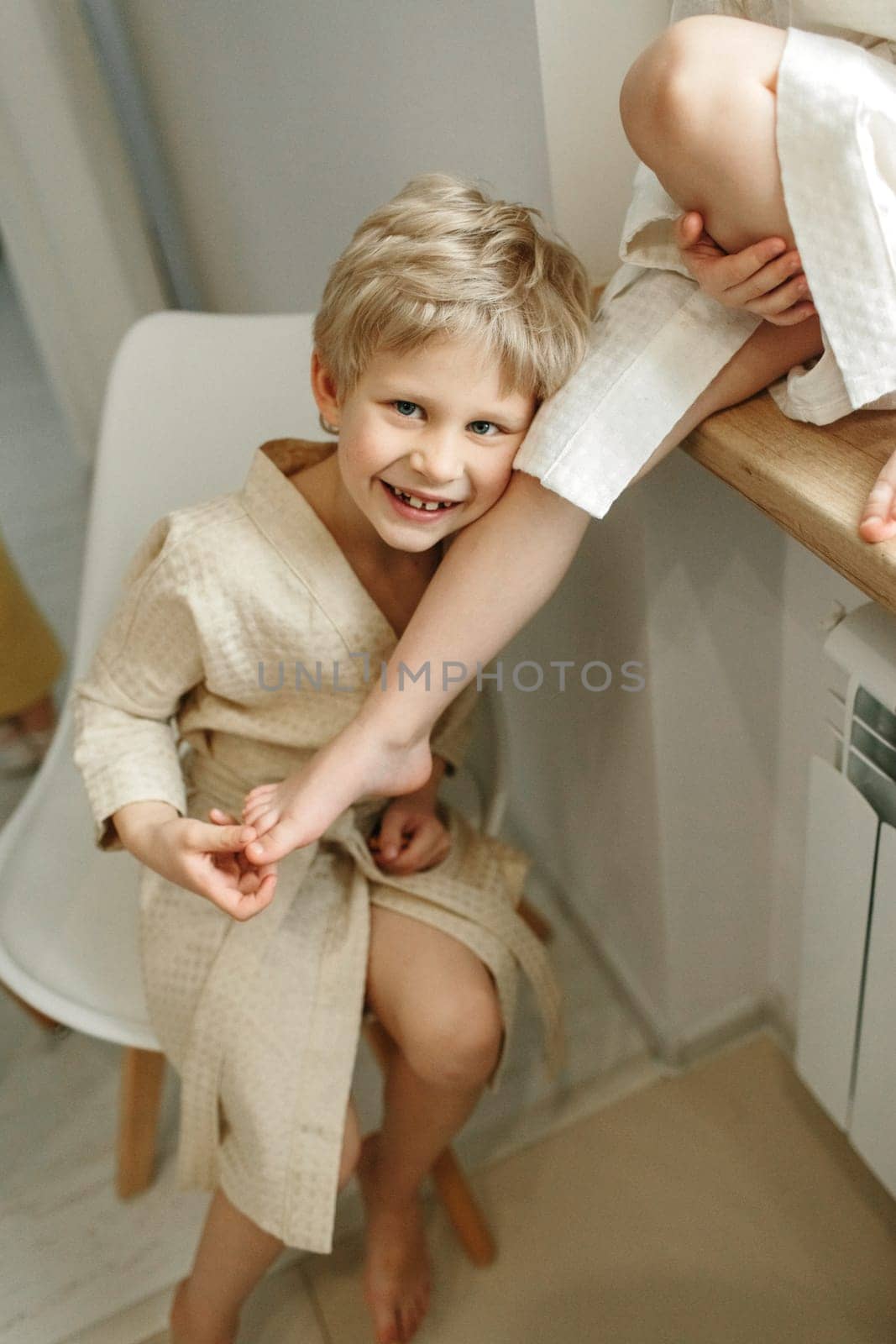 Portrait of a fair-haired smiling boy in a bathrobe who sits on a chair and leans on the girl's leg.
