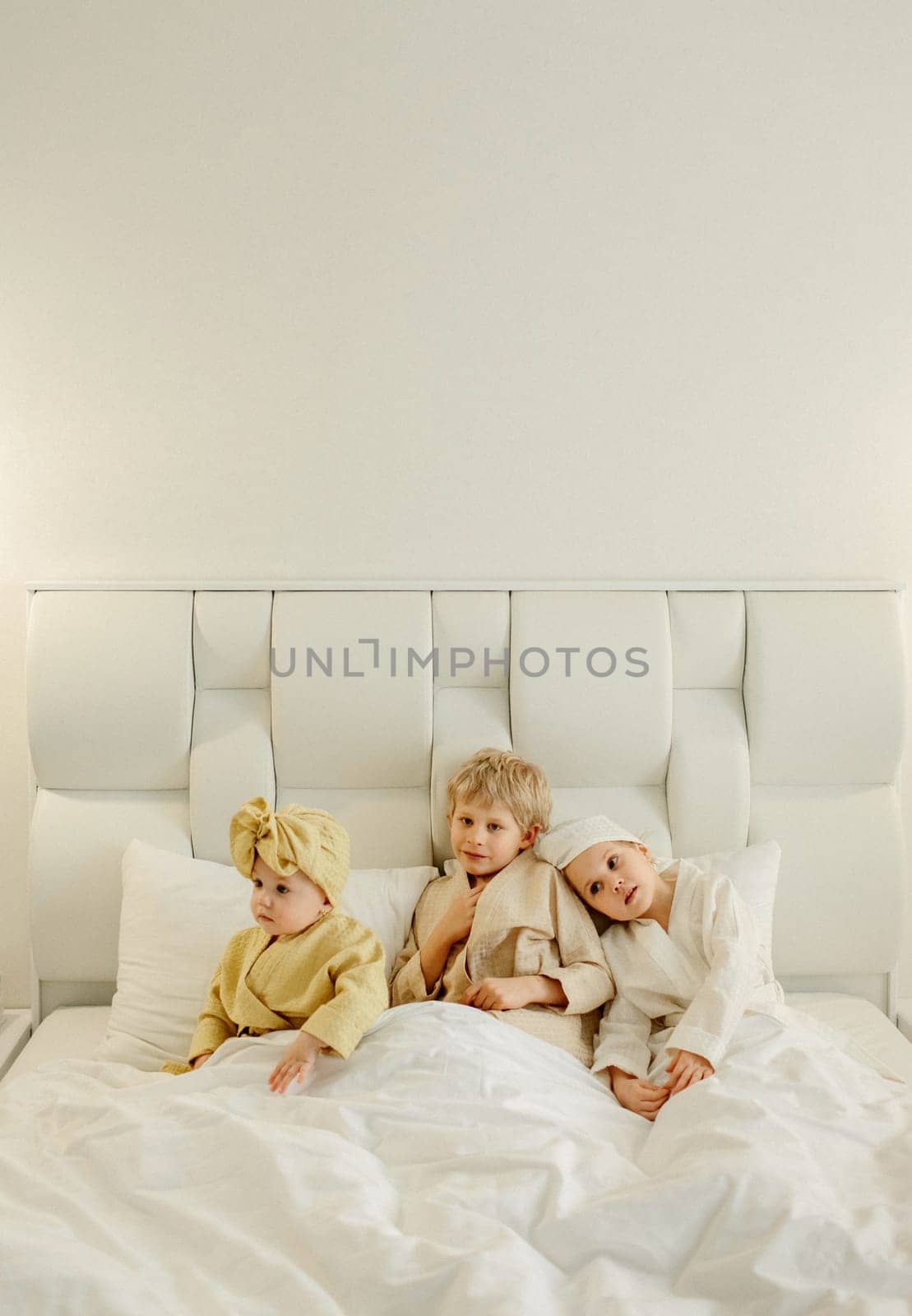 Children lie in a white bed in bathrobes after taking a bath, smiling and looking at the camera. View from above.