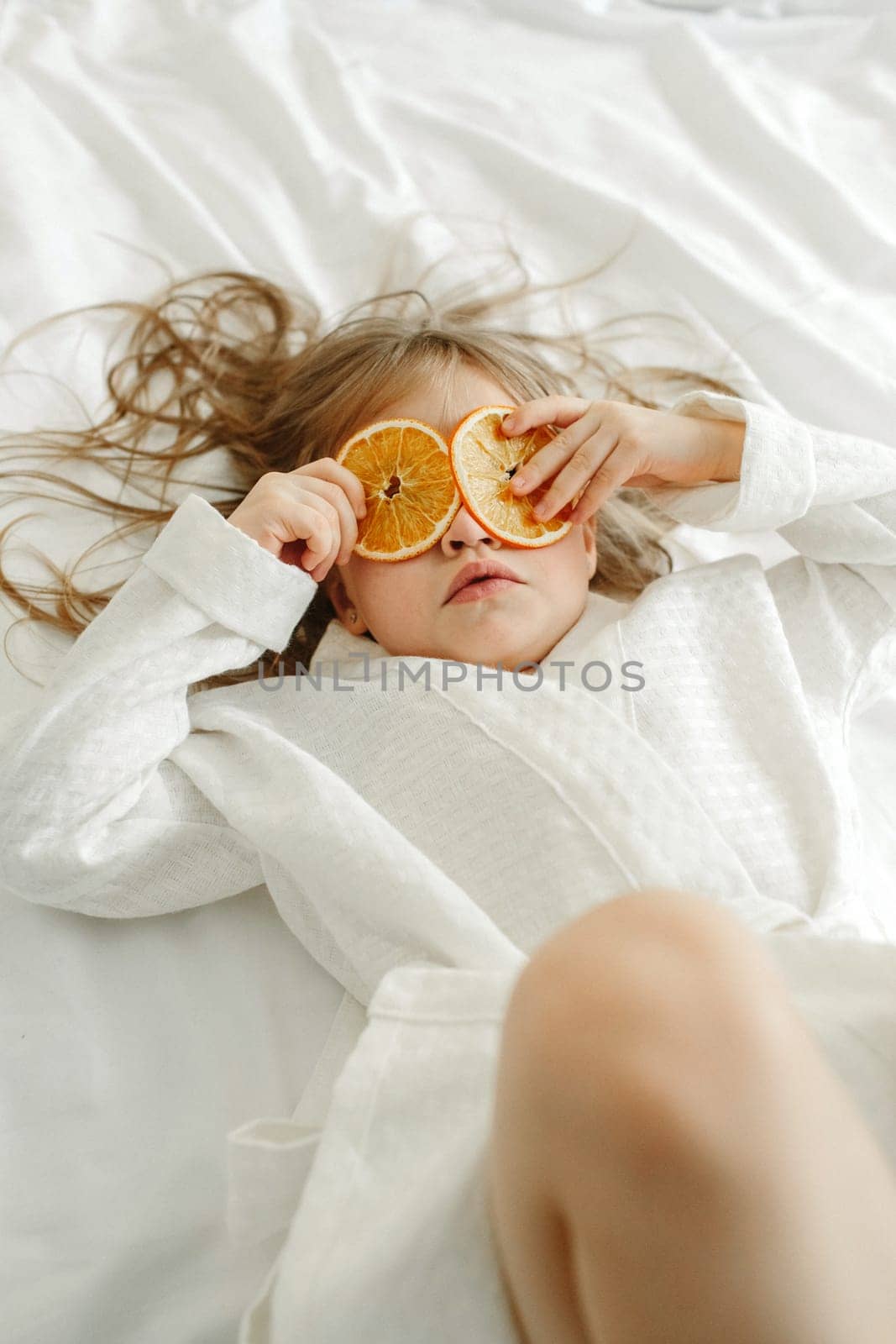 A young girl in a white coat, lying in bed, playing with candied oranges, closed her eyes with them.