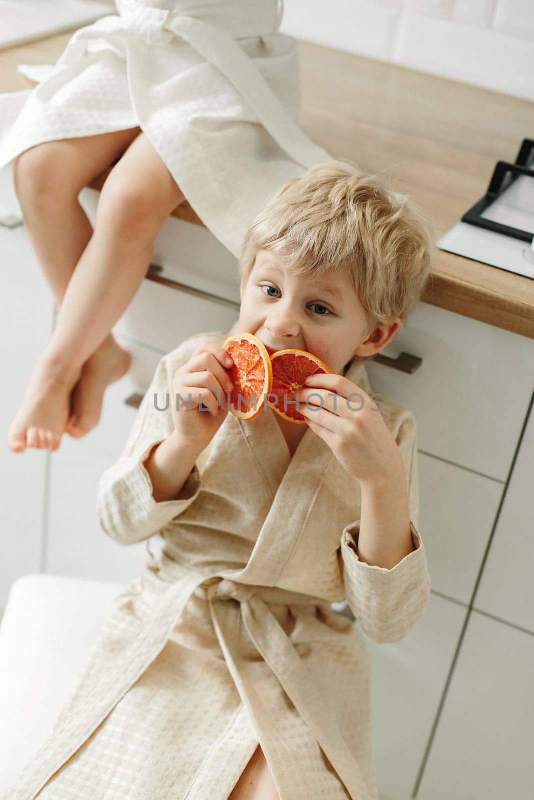 A boy in a bathrobe sits in the kitchen and holds candied oranges in his hands.