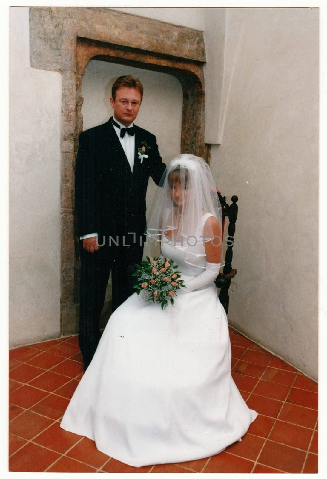 Retro photo shows newlyweds. Bride wears long veil, white delicate wedding gown and bridal gloves. She holds a small bouquetof roses. Vintage color photography. Circa 1990. by roman_nerud