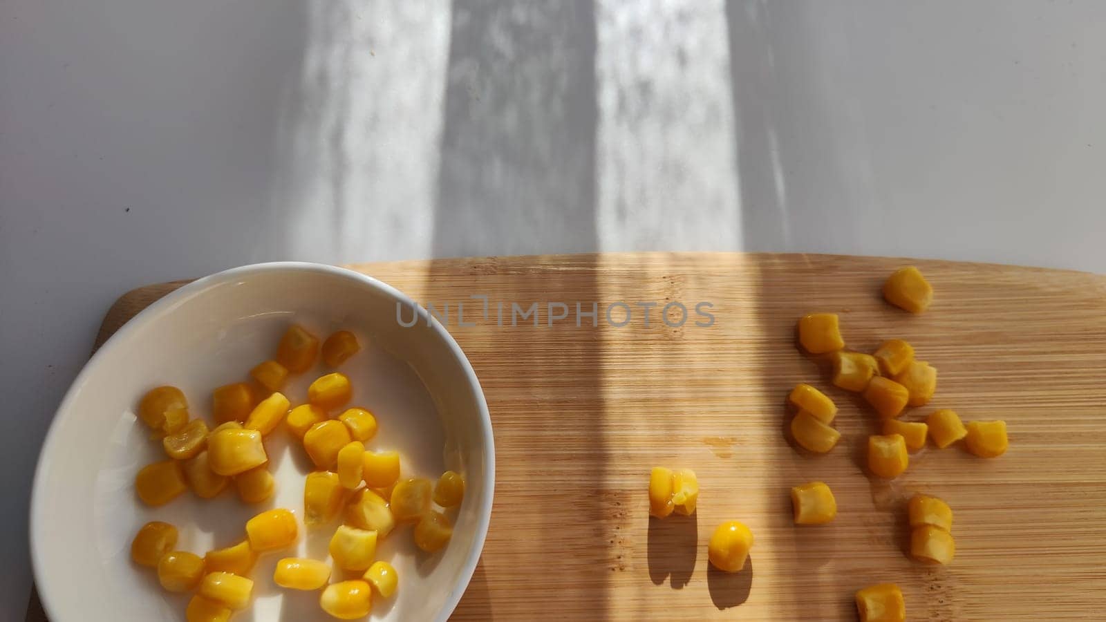 Lot of pieces of canned yellow corn on plate which is on wooden bamboo cutting board on white background. Concept of cooking and delicious healthy food