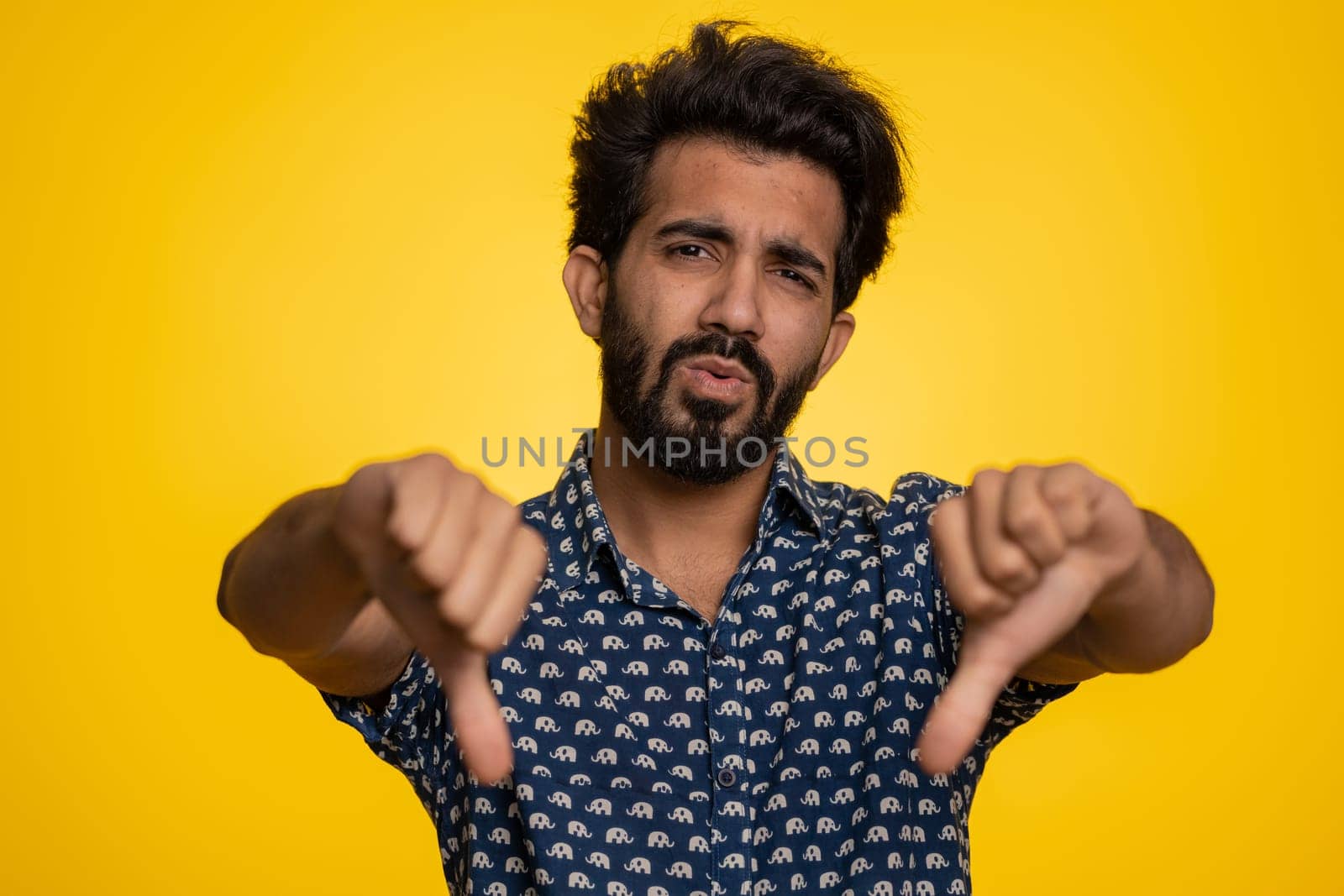 Dislike. Upset unhappy indian man in shirt showing thumbs down sign gesture, expressing discontent, disapproval, dissatisfied, dislike. Handsome young guy. Indoor studio shot on yellow background