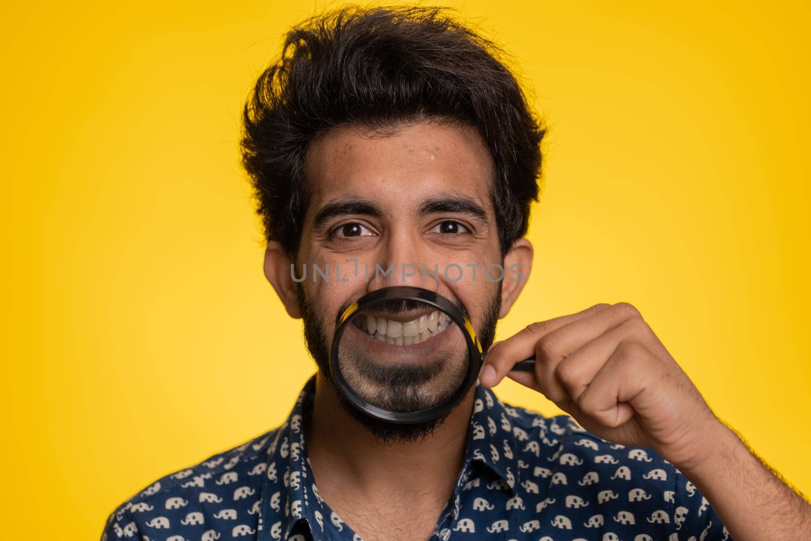 Indian man holding magnifier glass on healthy white teeth, looking at camera with happy expression, showing funny silly face smiling mouth. Handsome hindu guy isolated on yellow studio background